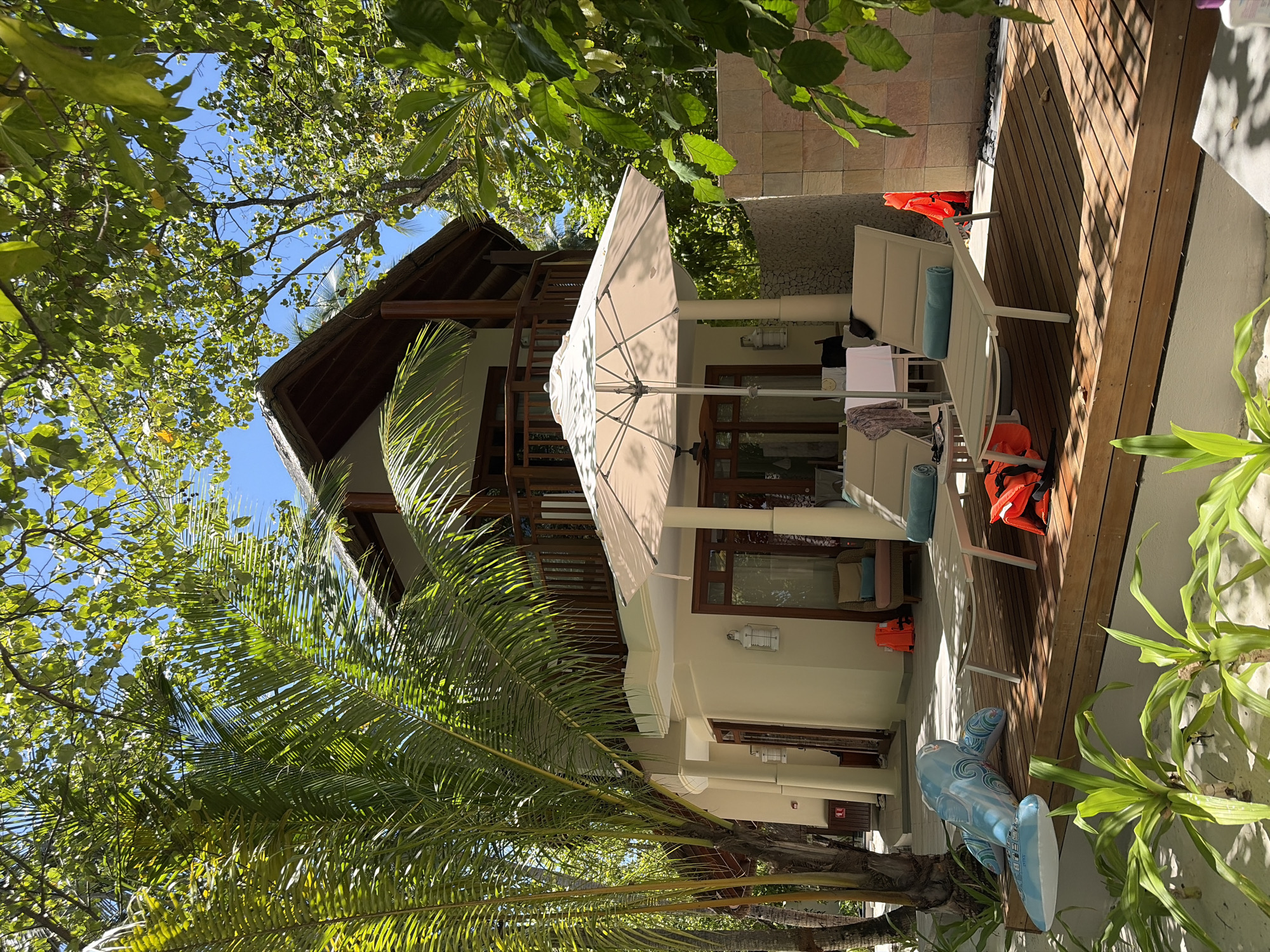 In the city of Halaveli, Maldives, a resort scene unfolds under a clear blue sky. A two-story building with a thatched roof and wooden balconies forms the backdrop. Lush green palm trees and foliage frame the structure, casting dappled shadows on the wooden deck in the foreground. Two lounge chairs with blue towels and orange life vests are arranged on the deck. An inflatable dolphin toy lies near the chairs, partially resting on the sand. An open umbrella shades a table set with chairs. The environment suggests a tropical vacation destination.