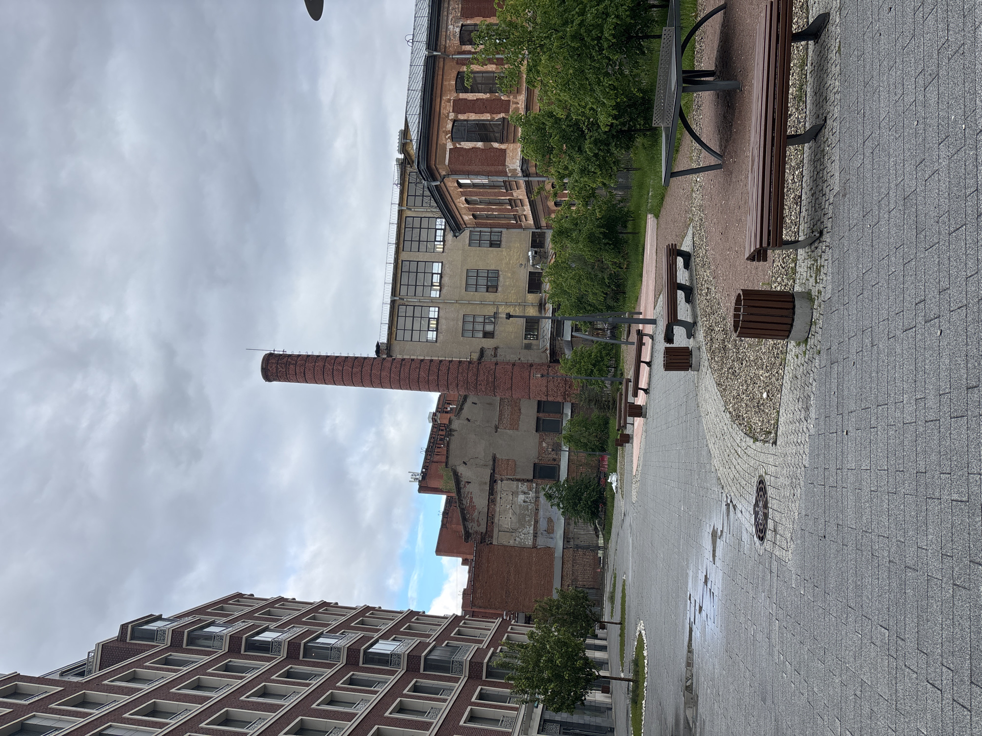 The image depicts a street scene in Saint Petersburg, Russia, under a cloudy sky. A tall, red brick smokestack rises prominently in the middle ground, suggesting a former industrial site. Adjacent to the smokestack is a collection of older brick buildings, some showing signs of decay or partial demolition.
In the foreground, a modern residential building with multiple floors and repetitive window patterns stands. A paved pedestrian walkway runs through the scene, featuring benches, trash receptacles, and small trees planted in regular intervals along the side. The pavement appears wet, indicating recent rain or damp conditions. The style of architecture and materials suggest a redeveloped or revitalized urban area, blending old industrial remnants with contemporary construction. There are no people visible in the scene.