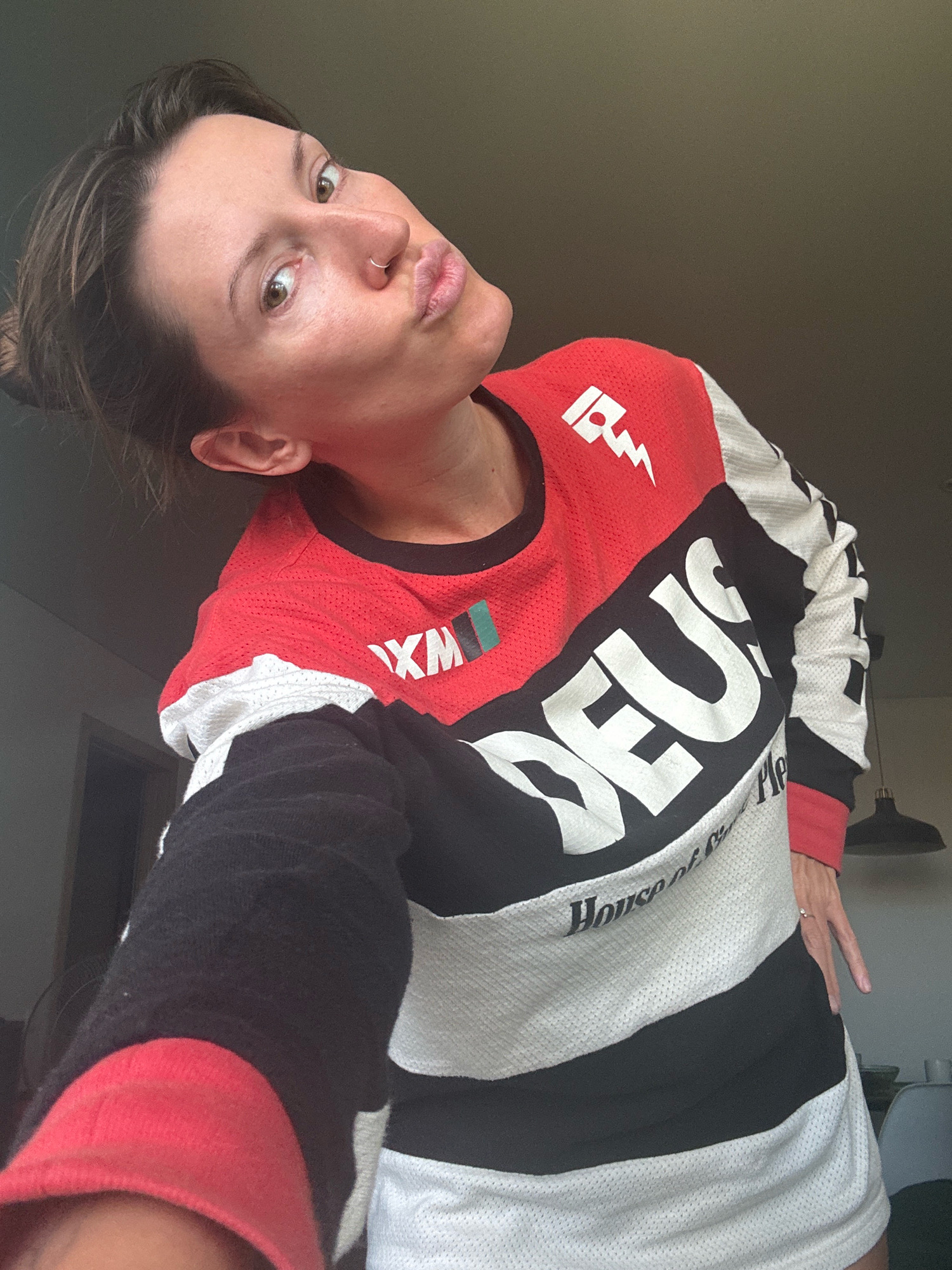A woman with brown hair and brown eyes, wearing a red, black, and white long-sleeved shirt with a "DEUS" logo, poses for a selfie. She has a nose ring and her lips are pursed. The background is a plain, dark wall. The image does not provide any information about the city of Bataysk, Russia.