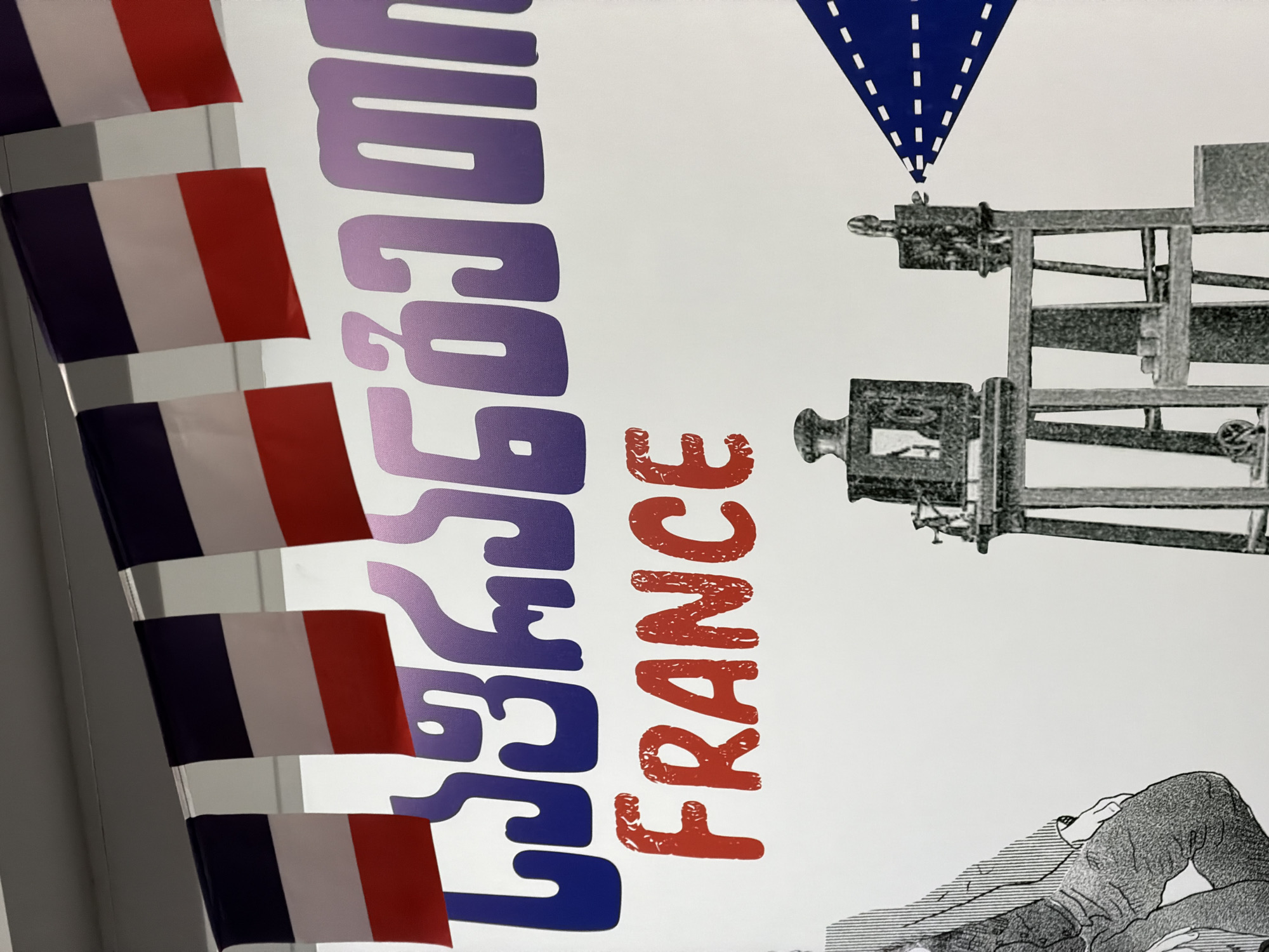The image depicts a poster or printed material, partially obscured by several small French flags. The poster features a combination of text and illustrations. The word "FRANCE" is prominently displayed in red. Next to it is an inscription in Georgian language. Above and to the right, there is a drawing of a vintage film camera, with a downward-pointing blue shape with white dashes resembling a stylized light cone. Below the camera is a sketched figure of a reclining person. The overall scene appears to be related to the presentation or promotion of French cinema or culture in Georgia. Without further context it's impossible to determine the exact location in Tbilisi.