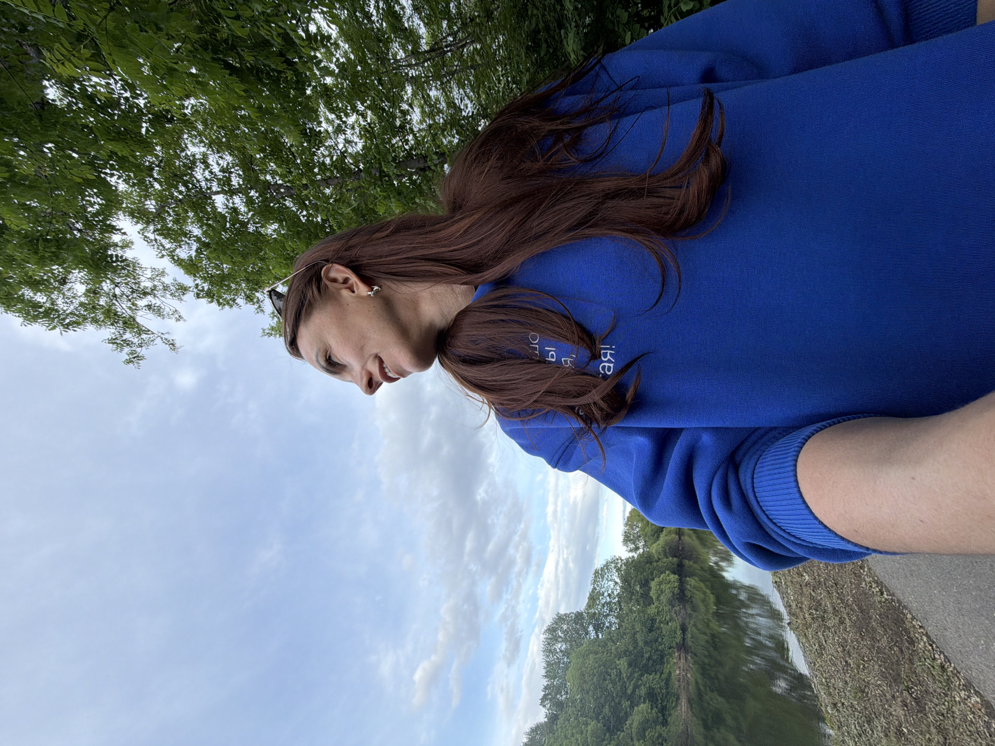 An outdoor, low-angle shot features a young woman with long, dark brown hair, standing in what appears to be a natural park setting in Birobidzhan, Russia. She is wearing a vibrant royal blue long-sleeved sweatshirt. Sunglasses are perched on top of her head, and a small, metallic earring is visible in her left ear. She has a subtle smile and appears to be looking towards her left.
The background consists of a partially cloudy, light blue sky above, and a lush green tree canopy on the upper left, suggesting an abundance of foliage. Below her, a body of water, likely a river or lake, stretches across the mid-ground, reflecting the trees and sky. On the far bank, more dense green trees line the water's edge. In the foreground, a paved or dirt path runs parallel to the water body.
The scene suggests a mild daytime, possibly overcast, providing soft natural lighting. The woman's relaxed expression indicates a pleasant or contemplative moment outdoors. On her blue sweatshirt, some white text is visible in reverse, appearing partially as "iRe" or similar lettering.