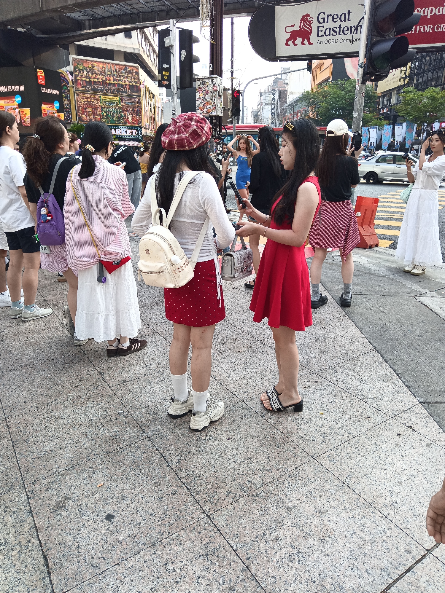The image shows a street scene in Kuala Lumpur, Malaysia, likely in a commercial or shopping district. Several young women are visible on a pedestrian walkway, standing near a crosswalk. One woman in the foreground, wearing a red dress and black sandals, is looking at her phone, while another woman next to her, in a red and white striped shirt and a white skirt, also holds a phone. A woman with a red and white checkered hat and a white backpack stands to the left, facing away from the camera. Other women, dressed in various casual outfits, are also present, some with backpacks and others looking at their phones. In the background, there are buildings with signage, traffic lights, and a glimpse of traffic. The ground is paved with granite tiles. The overall impression is a busy urban environment with people engaged in everyday activities.
