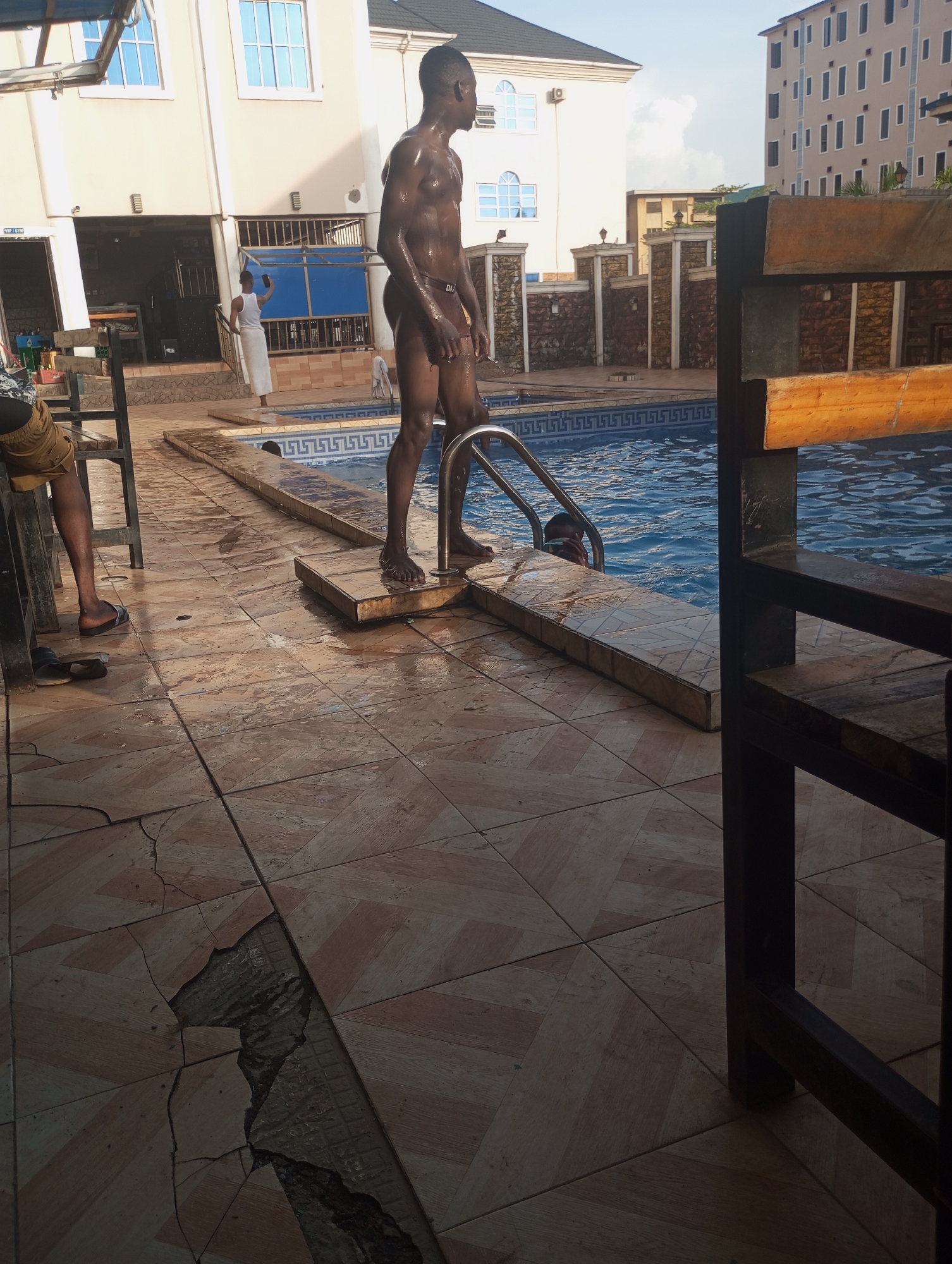 A man stands near a swimming pool next to a building in Onitsha, Nigeria. The man is dark-skinned and shirtless, wearing dark underwear. Another man in a white towel holds his phone up to take a photo. One person is in the pool. The scene is taken next to a partially broken tile.