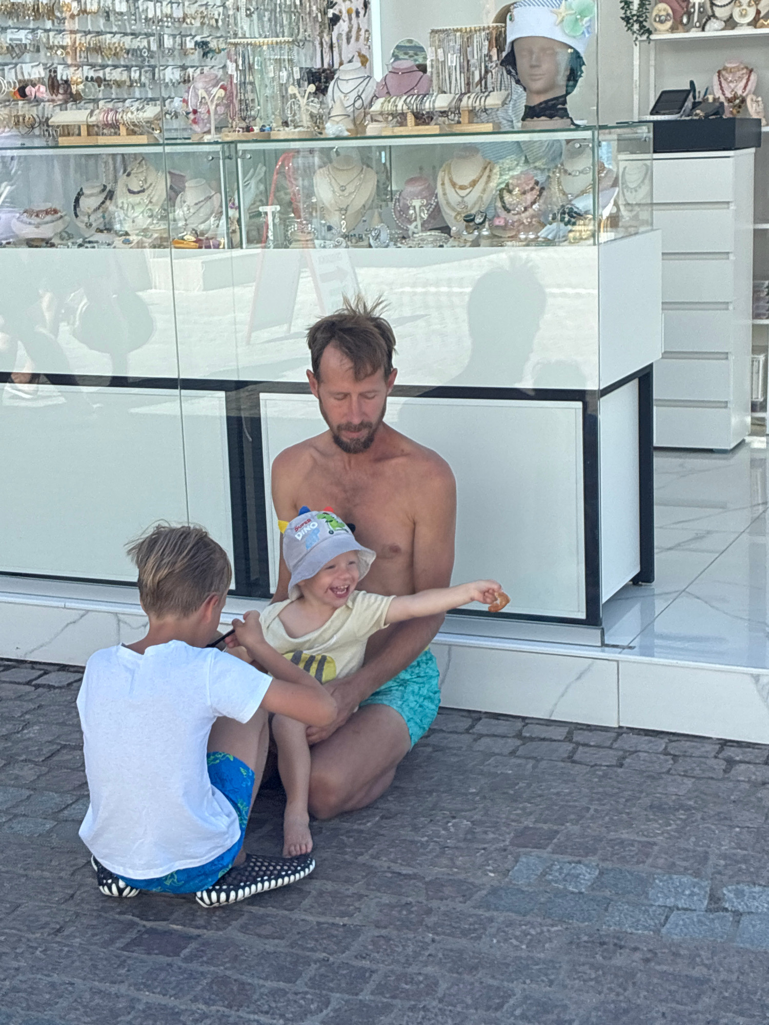 In Sochi, Russia, a man is kneeling on cobblestone pavement, holding a toddler on his lap. The toddler, wearing a light-colored bucket hat and a yellow t-shirt, is laughing and reaching out. Next to them, a young boy in a white t-shirt and blue swim trunks is also seated, looking down at the toddler. Behind them is a brightly lit display of jewelry and accessories in glass cases, suggesting the location is a shop or market. The scene appears to be outdoors, with a reflective surface in the background indicating either glass or a polished floor.