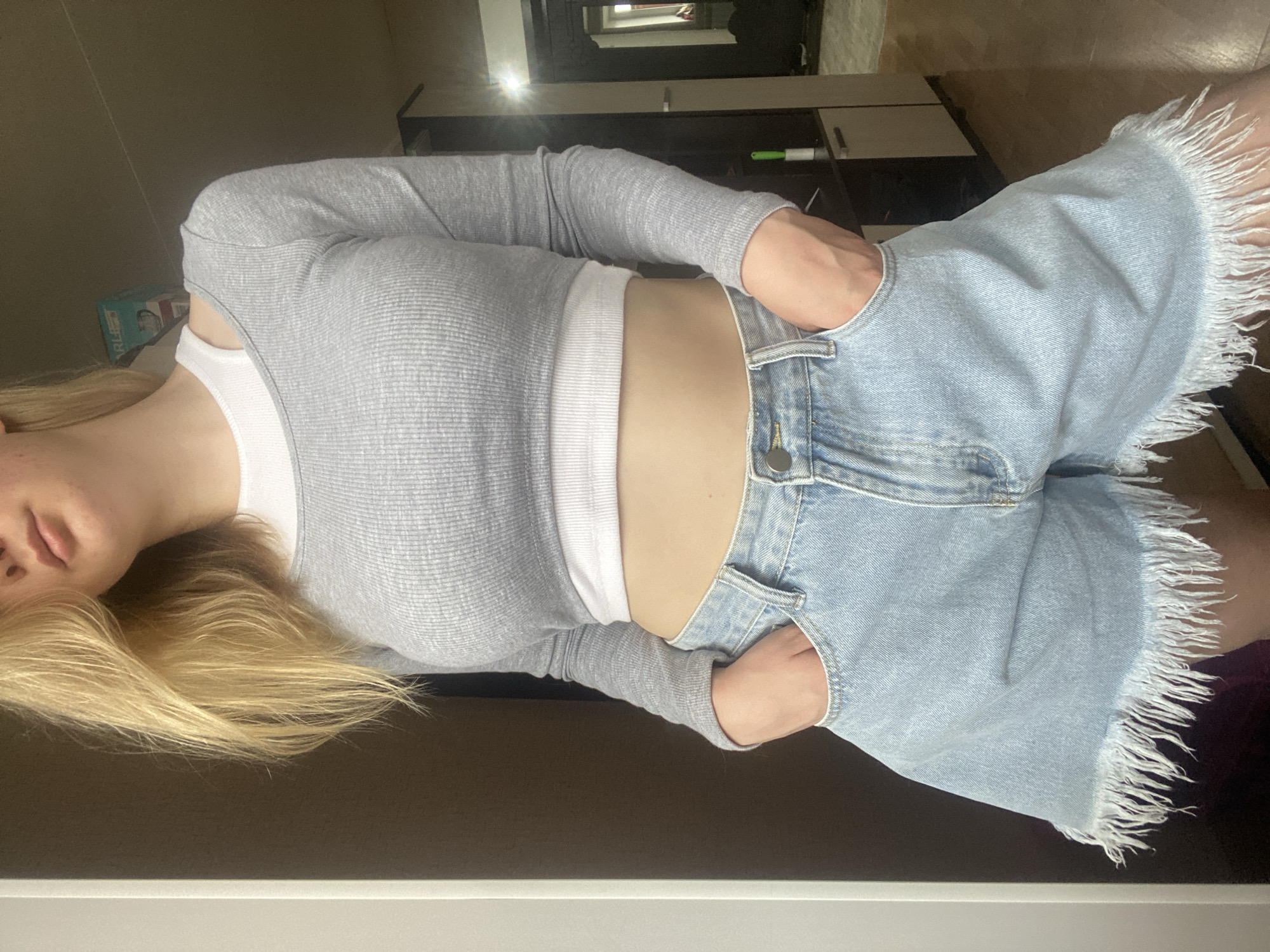 The image shows a person from the chest down, lying on their side with their head resting on their arm. They are wearing a grey long-sleeved ribbed top over a white tank top, and light blue denim shorts with a frayed hem. Their hands are tucked into the pockets of their shorts. The background appears to be an indoor setting, possibly a bedroom or living area, with wooden flooring and a dark wall. There is a glimpse of a shelf unit with some objects on it in the background, including what looks like a lint roller. The lighting suggests it might be daytime, with light coming from a window, casting shadows and creating reflections on the surface behind the person. The overall mood is relaxed and casual. No visible text is present in the image.