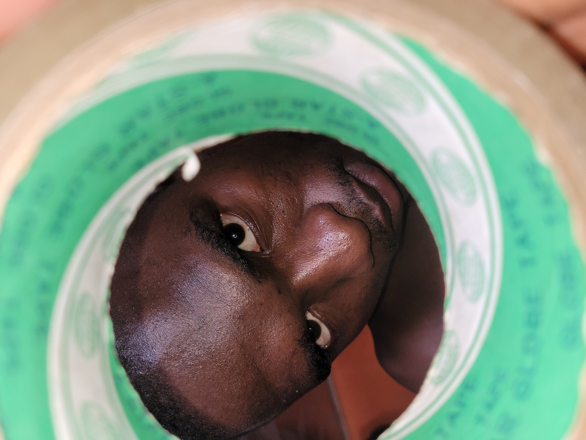 A close-up, low-angle, over-the-shoulder shot shows a Black man's face looking directly into the camera, peering through the center opening of a roll of clear packing tape. The man, whose face is partially obscured by the green and white edges of the tape, has dark skin, short black hair, and thick eyebrows. His left eye is wide open and focused on the lens, while his right eye is slightly narrowed. He has a faint mustache and a small patch of stubble on his chin. The tape roll has green printing on its inner surface, with blurry white lettering that appears to read "GLOBE TAPE." The background is out of focus, with an orange surface visible behind the man's left shoulder. The image appears to have been taken indoors, possibly in Bukuru, Nigeria.