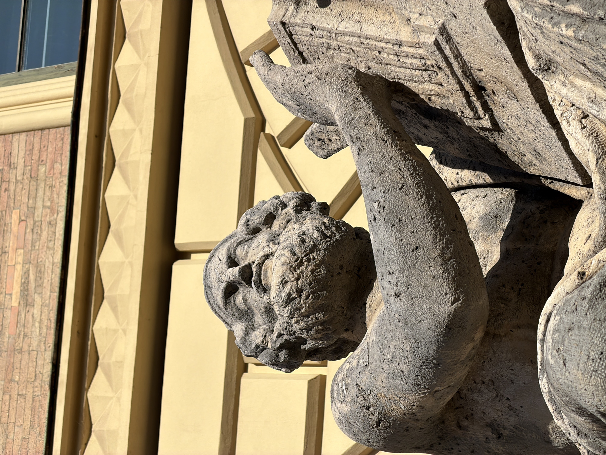 The image shows a close-up, low-angle view of a weathered stone sculpture of a man with a beard, seated and holding a book or tablet. The sculpture is made of rough, grey stone with visible pockmarks and cracks, indicating age and exposure to the elements. The man has his eyes closed or downcast, and his facial features are worn. His right arm is raised, and his hand rests on the object he is holding. The sculpture is positioned in front of a building with a pale yellow facade, which features a decorative band with a geometric pattern. Above the yellow facade, a brick wall can be seen. The scene is outdoors and bathed in bright sunlight, casting distinct shadows on the sculpture and the building. The context of this sculpture is likely architectural decoration or a public monument, given its scale and placement against a building. This sculpture is part of the Elias-Holl-Denkmal (Elias Holl Monument) in Augsburg, Germany, not Munich.