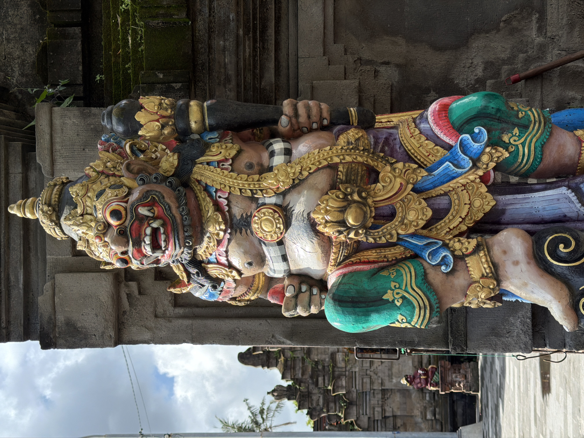 A detailed image depicts a Balinese statue of a deity, possibly a guardian figure, located in North Kuta, Indonesia. The statue is horizontal and displays intricate carvings and vibrant colors, including gold ornamentation, red facial features, green clothing, and blue accents. It has a fierce expression with exposed teeth. The statue holds a black club. The background features weathered stone architecture, some covered in moss, suggesting an ancient temple or site. A glimpse of the sky and a palm tree can be seen at the bottom. A second, small statue is visible in the background.