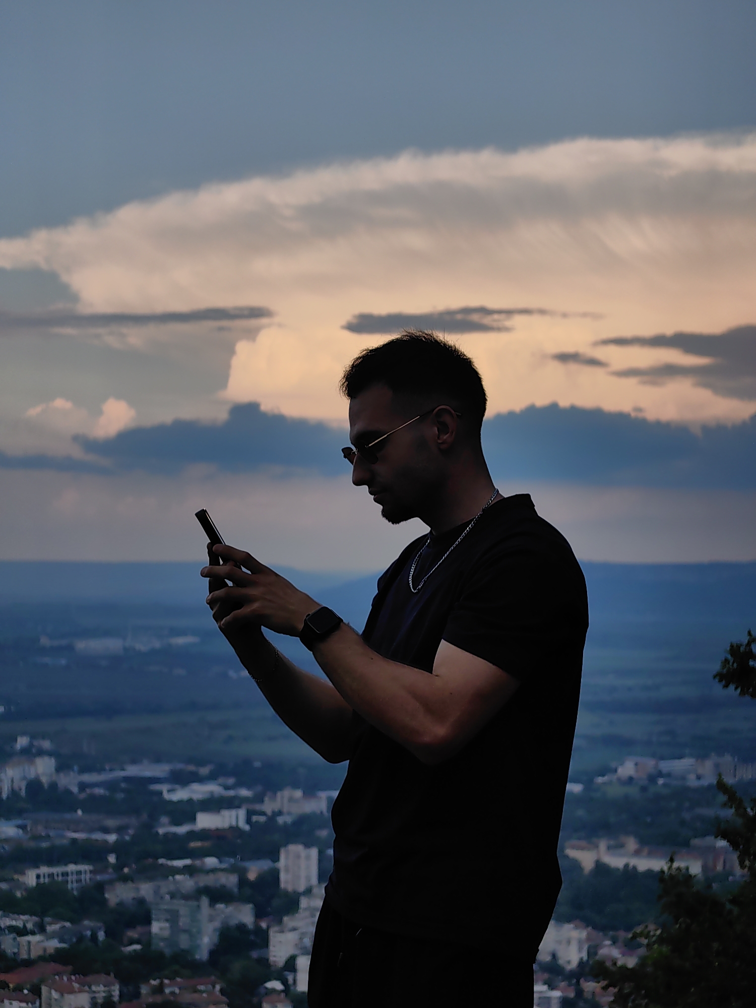 A medium shot depicts a man in silhouette using a smartphone with a cityscape in the background. The man is viewed from the side, looking down at the phone. He is wearing sunglasses, a black t-shirt, a chain, and a smartwatch. The background reveals a cloudy sunset sky above a distant cityscape. Based on the landmarks and urban layout visible, the location is likely near Shumen, Bulgaria, potentially taken from a higher vantage point overlooking the city. The man is likely taking a photo or interacting with the phone while enjoying the view.