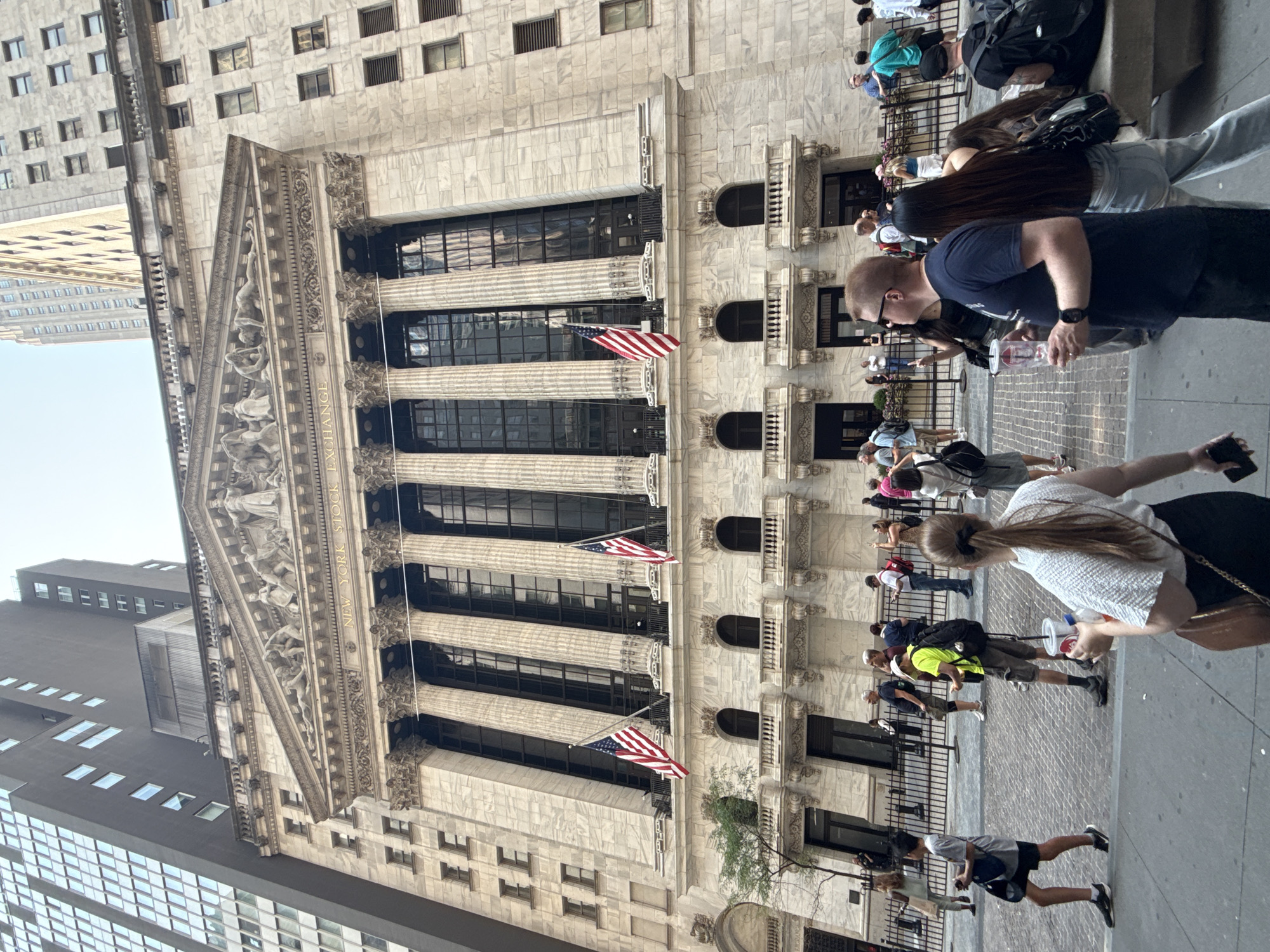 The image shows the New York Stock Exchange building in New York City, United States of America. The iconic neoclassical facade of the building, characterized by its large Corinthian columns and intricate sculptures above the entrance, is prominently displayed. Two American flags are visible, one on each side of the central section of the building.
In the foreground, a diverse group of people are walking on the sidewalk. Many are tourists, some holding drinks and taking photos. A young boy in a gray t-shirt and black shorts is walking towards the left, carrying a blue bag. A man in a neon yellow t-shirt and shorts, wearing a backpack, is walking towards the right, looking at his phone. A woman with long brown hair tied in a ponytail, wearing a white patterned top and black skirt, is walking towards the foreground, holding a cup and a bag. Another man in a dark blue t-shirt and sunglasses is walking in the middle ground, holding a clear cup.
The street is paved with cobblestones. Behind the people, a black metal fence separates the sidewalk from the building's entrance. In the background, modern skyscrapers rise above the historic stock exchange building, indicating the urban environment of New York City. The sky is overcast.