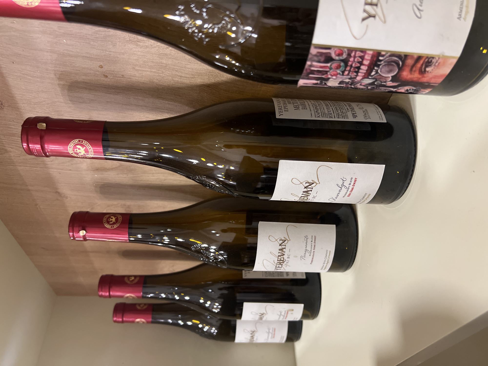 The image shows a selection of "Yerevan" brand wine bottles. They are lying horizontally on a light-colored surface with a wooden background. The bottles are of different sizes, with red foil caps. Their labels indicate "Pomegranate Wine" and "Winemakers blend", produced by "Artenia Wine Company", and the "Karmirayut" wine, whose Russian text describes the place of production and the alcohol volume in Russian language. Yerevan city and Armenia are visible on the labels.