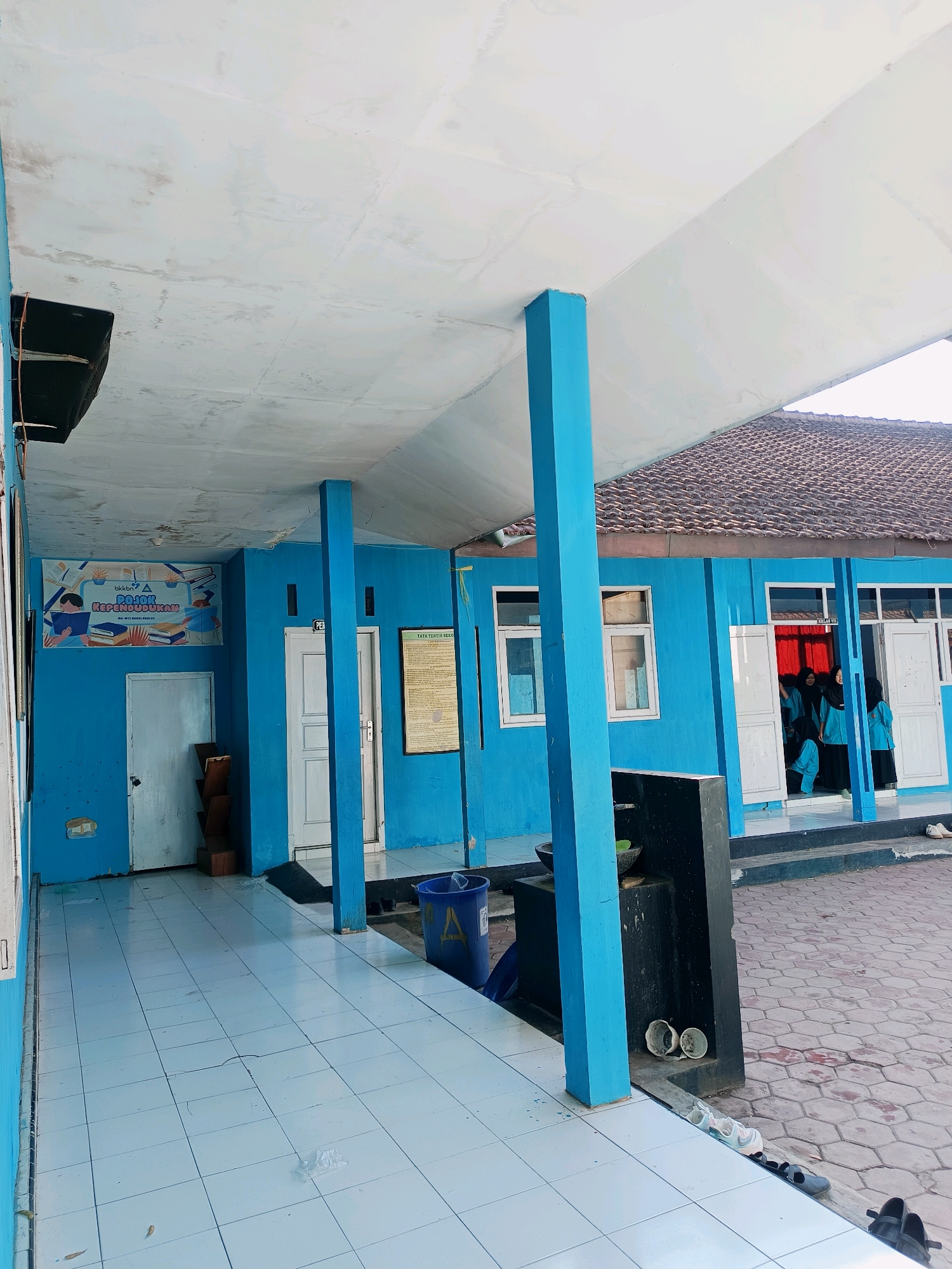 The image depicts the exterior of a blue building, likely a school or public institution, in Panggungan, Indonesia. A covered walkway with a white ceiling and blue pillars runs along the front of the building. The walkway is paved with light blue square tiles, some of which appear cracked or damaged. Several doors and windows are visible on the blue building. To the left, a poster with the text "BKKBN" and an image of a person reading is displayed on the wall. A wooden shelf with books is positioned near a white door. Further down the walkway, a blue trash bin is visible. On the right side of the image, an open doorway reveals several individuals in blue uniforms and headscarves, presumably students, standing inside. The ground outside the walkway is paved with hexagonal stones. Various objects are scattered around, including shoes near the entrance, and what appear to be ceramic bowls. The overall scene suggests a functional, perhaps slightly worn, educational or community facility.