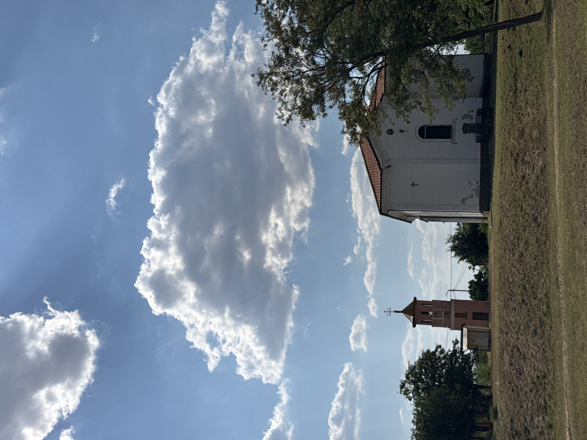 The image depicts a rural landscape with two structures that appear to be religious buildings. On the left is a tall, slender bell tower, painted a muted reddish-pink hue, topped with a cross. Adjacent to its base is a small, utilitarian structure with a white door. To the right of the bell tower, and closer to the foreground, stands a white building with a red-tiled roof, likely a church, featuring a cross on its facade and a barred window. A mature tree with lush green foliage leans into the frame from the right, partially obscuring the church. The ground in the foreground is covered in dry grass, suggesting a warm, possibly summer, climate. The sky is a vivid blue, populated by dramatic, fluffy white clouds that cast shadows. The scene evokes a sense of tranquility and historical significance, typical of a village setting in Bulgaria. The location in Ekzarh Antimovo, Bulgaria, is consistent with the architectural style and rural environment.