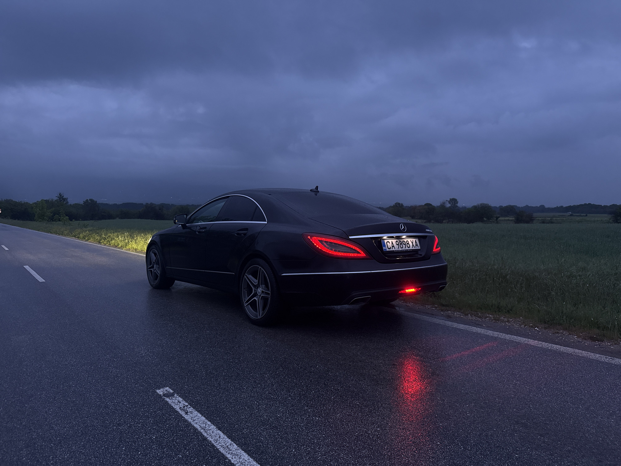 The image depicts a black Mercedes-Benz CLS parked on a wet asphalt road. The car's taillights and left reverse light are illuminated. The license plate reads "CA 9898 XA." The road has a white line marking its edge. Adjacent to the road is a field with green vegetation. In the background, a dark, overcast sky dominates the scene, with hints of trees and distant hills. The location is inferred to be near Kliment, Bulgaria, based on the license plate code.
