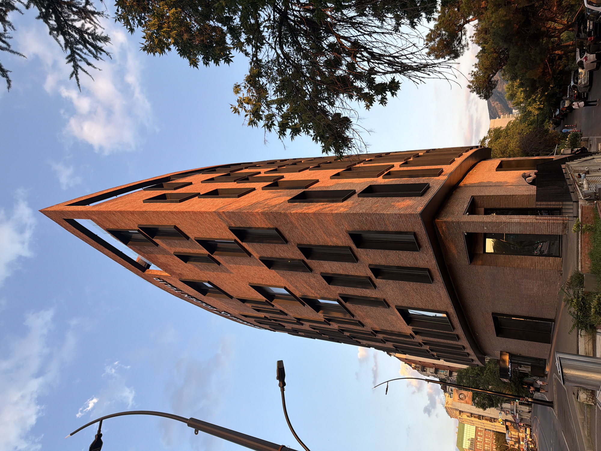 An eye-level, wide shot captures the unique architecture of the Golden Tulip Hotel, a brick building with a distinctive triangular shape, on a street in Tbilisi, Georgia. The building's facade is illuminated by the warm glow of the setting sun, highlighting the texture of the bricks and the dark frames of its numerous rectangular windows. Above the windows, the hotel's name, "GOLDEN TULIP," is visible in white lettering. A tall, dark lamppost with a modern design stands on the left side of the frame, its curved arm reaching towards the sky.
In the foreground, to the left of the hotel, a metal trash receptacle is situated on a paved area. A street runs along the base of the building, with a sidewalk bordering it. A few pedestrians, including two individuals walking together and one person standing on a balcony to the right, are visible. On the right side of the frame, lush green trees with sparse branches overhang the scene, their leaves catching the sunlight. Beyond the trees, a narrow street is lined with parked cars, and in the distance, a mountain range is silhouetted against the sky, bathed in the soft light of dusk. The sky above is a clear blue, with a few wispy clouds.