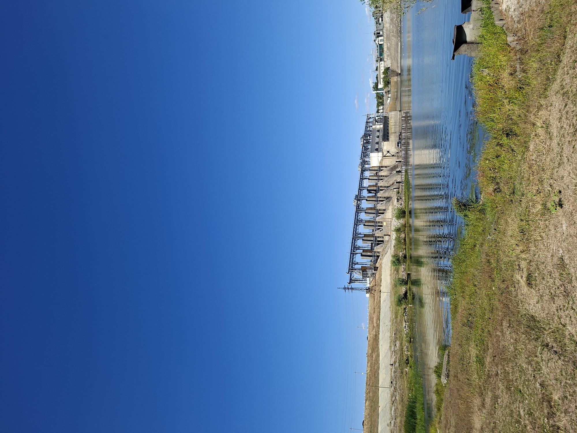 The image shows a wide shot of the Dubasari Dam and Hydroelectric Power Station on the Dniester River, located in Ustia, Moldova. The sky is a clear, bright blue. The dam structure is prominent in the mid-ground, a long concrete and metal edifice with multiple spillways and a building complex. Electrical towers and lines are visible on the embankment behind the dam. The river in the foreground is calm and reflects the blue sky and the dam. The riverbank on the left is covered in dry, golden-brown grass and some patches of green vegetation. There are no people visible in the image. The scene is serene and captures the industrial infrastructure integrated with the natural landscape.