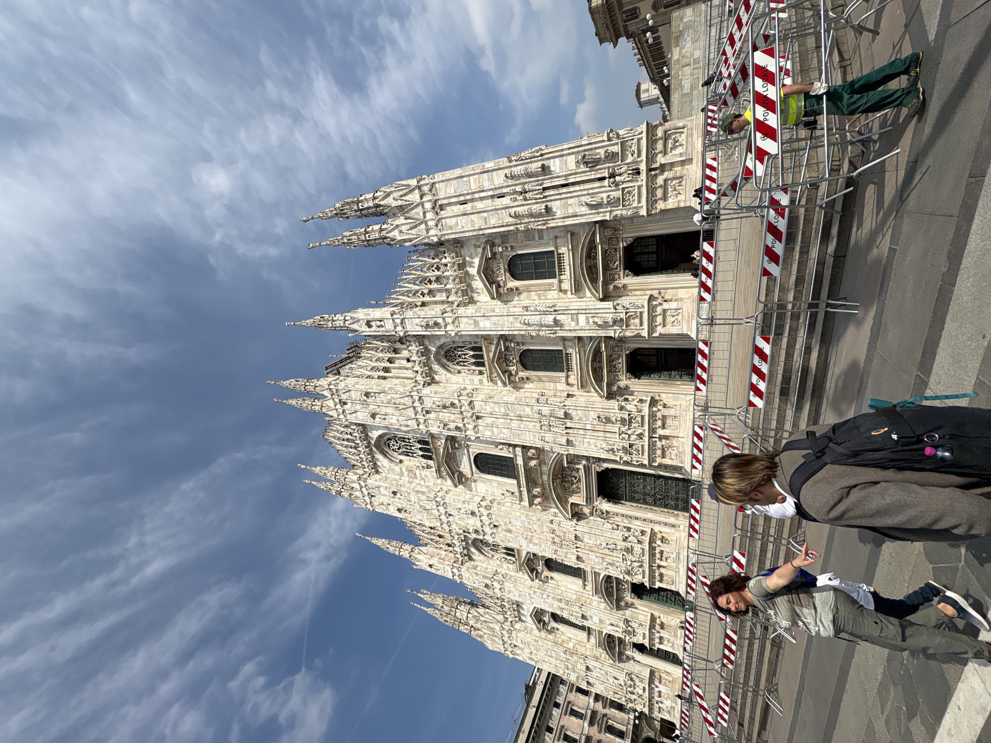 The image, rotated to display correctly, presents a vibrant daytime scene outside the iconic Milan Cathedral (Duomo di Milano) in Milan, Italy. The magnificent Gothic cathedral, crafted from white marble, dominates the left side of the frame with its incredibly intricate facade, numerous spires, ornate statues, and detailed architectural carvings.
In the foreground on the right, several people are present in what appears to be a public square or plaza. Two individuals are prominent: one with light brown hair, wearing a dark jacket over a white top, a white face mask, and a black backpack with a pink water bottle, is seen from the side. Beside them, another person with darker, curly hair, dressed in a gray t-shirt and cargo pants, is captured mid-gesture with their hand.
Further back, a worker is visible behind a series of red and white striped metal crowd control barriers. This person wears a high-visibility yellow-green vest, green pants, and a cap. The barriers, stretching across the mid-ground, feature the prominent text "POLIZIA LOCALE," indicating the presence of local police or official event management. The sky above is a clear blue with scattered wispy white clouds. The overall scene captures the grandeur of the cathedral amidst daily activity and crowd management.