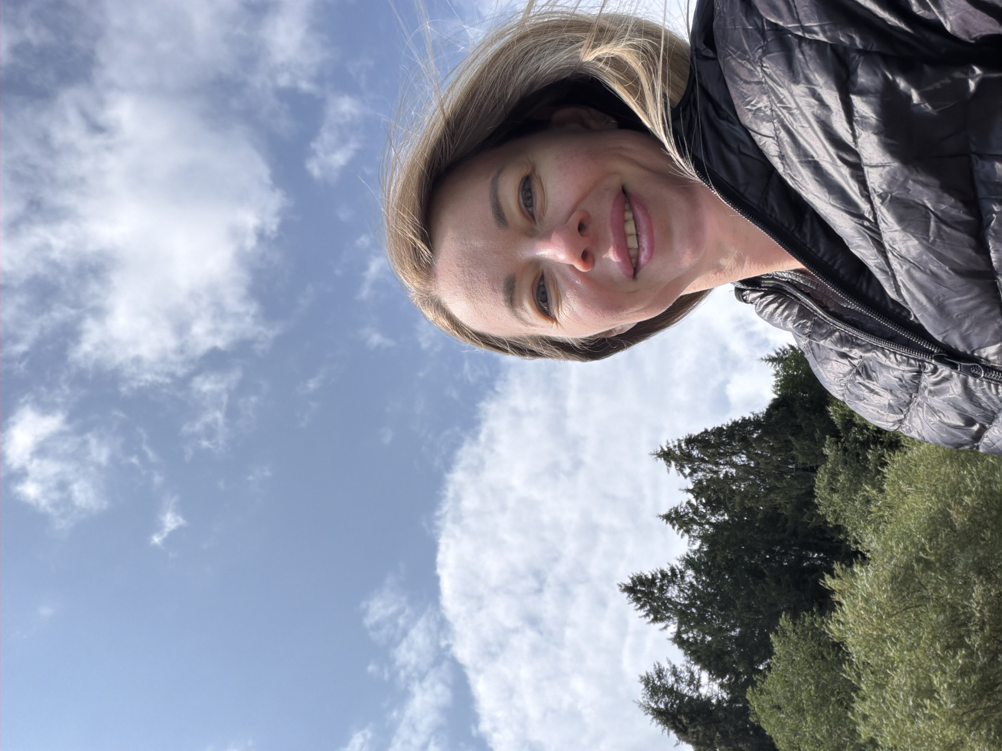 A woman with blonde hair and blue eyes is taking a selfie outdoors. She is wearing a black puffer jacket. The background shows a blue sky with white clouds and a line of green trees. The image is a close-up, with the woman's face and shoulders filling most of the frame.