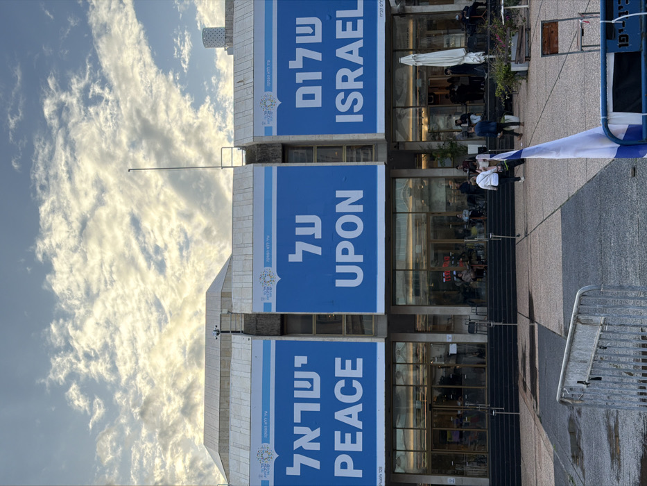 An eye-level photograph captures the Tel Aviv-Yafo Municipality building, located in Rabin Square, Tel Aviv, Israel, under a partly cloudy sky. The building's facade is clad in light-colored stone, featuring large glass windows and multiple entrances at ground level, accessible via dark staircases.
Three prominent blue banners are affixed to the building's upper facade. From left to right, these banners display Hebrew text "ישראל" (Yisrael), "על" (Al), and "שלום" (Shalom), with corresponding English translations "PEACE," "UPON," and "ISRAEL" directly below them. The complete message reads "שלום על ישראל" / "PEACE UPON ISRAEL." Small text on the bottom left of the leftmost banner reads "Psalm 128," while similar Hebrew text "תהילים קכח" is on the rightmost banner. Each banner also features a circular emblem at its top. A thin, dark pole extends vertically from the rooftop between the left and middle banners; security cameras are visible on the roofline above the left banner.
The foreground consists of a paved public square with a mosaic pattern of light and dark grey tiles. A silver-colored metal crowd control barrier is positioned in the lower-left. Partially visible in the lower-right, behind a similar barrier, is an Israeli flag, white with two horizontal blue stripes and a central blue Star of David, attached to a pole.
Numerous individuals are present within the scene. Several people are walking across the square in the middle ground, including one person in a light-colored top and dark pants moving right, and another in a dark jacket behind them. Others are visible near the building's entrance and seated at outdoor tables on the right side, where a large white patio umbrella is open. Through the building's glass facade, additional individuals are discernible inside. Potted plants are positioned near the building's base. The overall lighting suggests late afternoon or early morning, with sunlight illuminating the undersides of the scattered clouds.