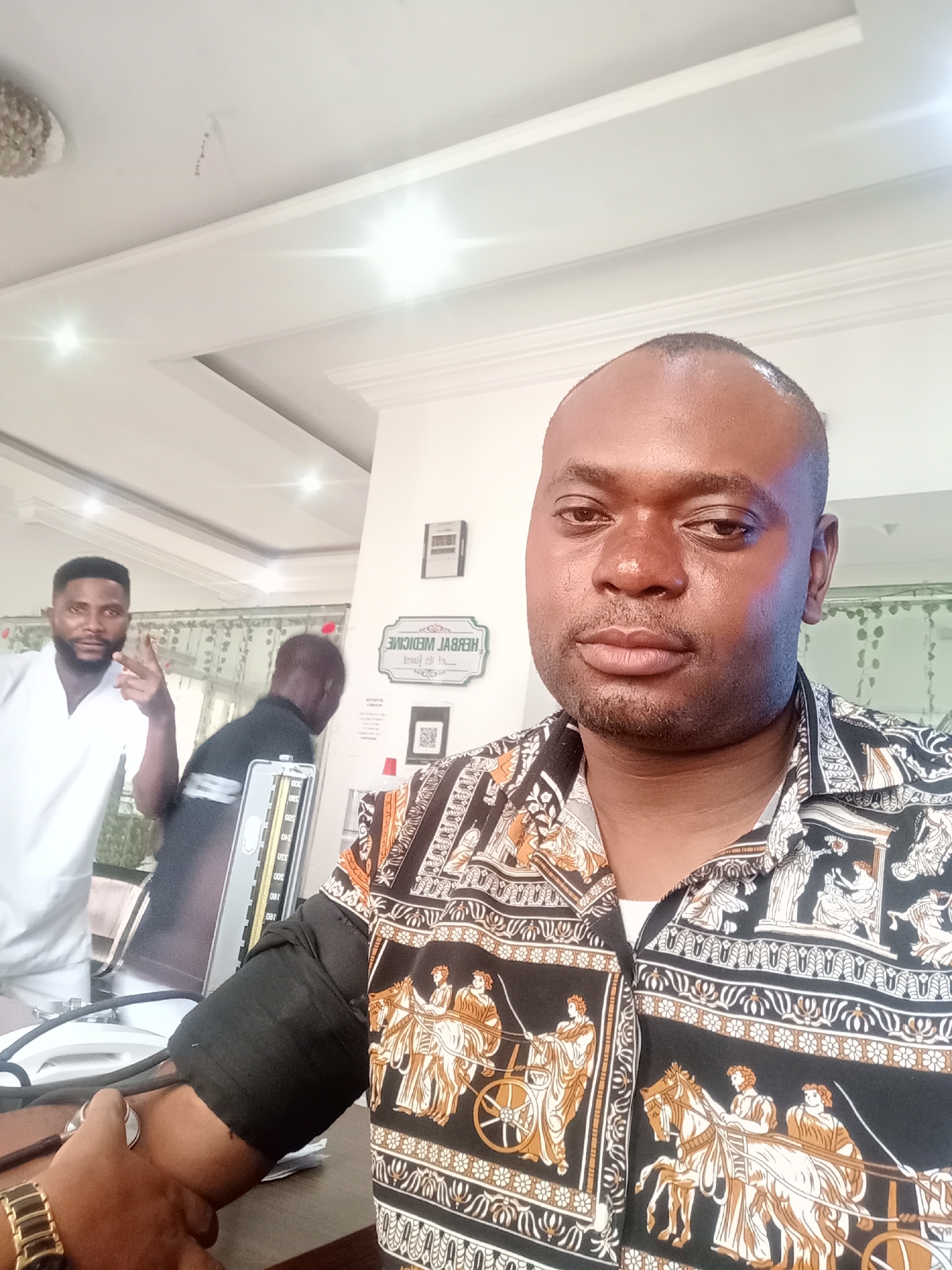 A man takes a selfie indoors. He wears a black and gold patterned shirt, a gold watch is visible on his left wrist as he appears to have his blood pressure taken. In the background, another man in a white coat holds up two fingers. A blood pressure measuring device is in the middle ground. The name “HERBAL MEDICINE” is on a plaque visible in the background. The location is Gwarinpa, Nigeria.