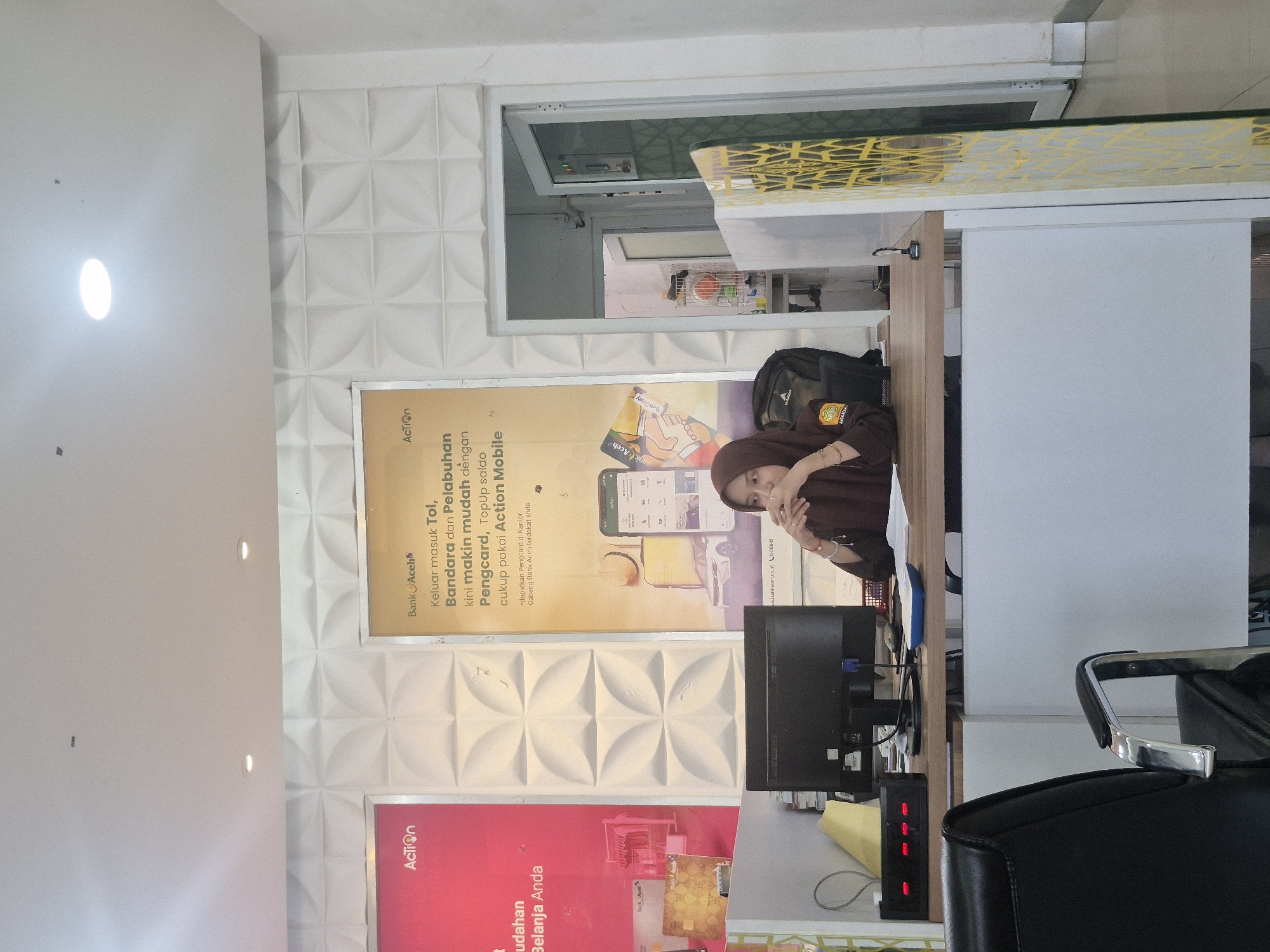 Here is a description of the image:
The photograph captures the interior of what appears to be a bank or financial institution, possibly a branch of "Bank Aceh" in Lhokseumawe, Indonesia. The room is brightly lit with a textured white wall featuring an advertisement for "Action Mobile" alongside the bank's logo. The poster promotes an electronic payment system for tolls, airports, and harbors, suggesting ease of use via mobile application and "Pengcard" payment card.
Behind a counter, an individual wearing a brown hijab sits at a desk with a computer monitor. There's a black backpack resting behind her, and a security or access control panel with red lights is visible near the monitor. The desk is cluttered with various items. The setting suggests an office or customer service environment within the financial institution. The picture also gives a partial view of a chair and the interior design, including a hallway or opening towards the rear.