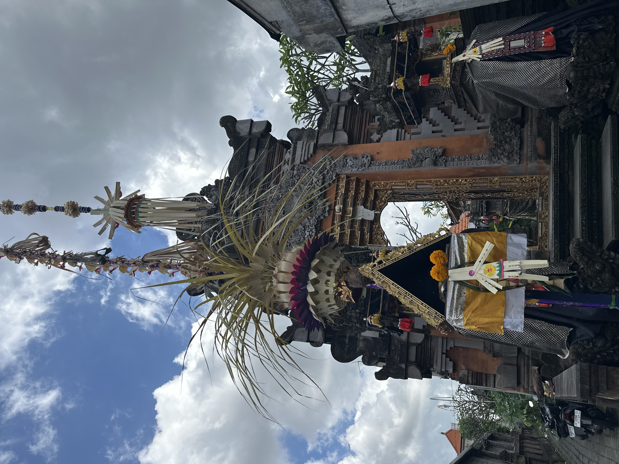 The image shows a Balinese temple entrance in Ubud District, Indonesia. The temple features traditional Balinese architecture with ornate stone carvings and a tiered roof. The entrance is decorated with festive offerings, including tall bamboo poles adorned with woven palm leaves, flowers, and colorful fabric. A mirror is incorporated into the entryway. In the foreground, a stone pathway leads up to the temple steps, with a parked motorcycle on the left. The sky is partly cloudy.