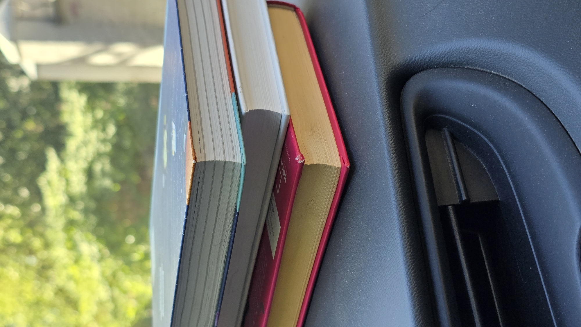 The image shows a close-up view of three books stacked on a dark, textured surface, possibly a car dashboard. The books are of varying sizes and colors. The top book has a dark blue cover with an orange and white pattern. The middle book has a white cover. The bottom book has a bright pink cover and a yellowed fore-edge. In the background, out of focus, is lush green foliage and a blurry light-colored structure. The foreground features a dark, ribbed air vent. No people or animals are visible in the image. The scene appears to be outdoors on a sunny day, given the lighting and dappled shadows.