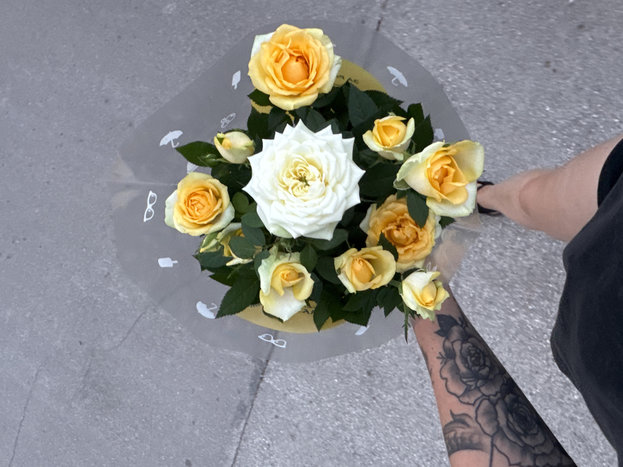 A bouquet of yellow and white roses is held by a person whose arm, adorned with a floral tattoo, is visible. The bouquet is wrapped in clear cellophane with a subtle pattern of white umbrellas and glasses. The roses are a mix of fully bloomed and budded flowers, with vibrant green leaves. The bouquet is resting on a gray, textured surface that appears to be pavement or asphalt. In the background, a portion of the person's legs and torso, dressed in dark clothing, is visible, suggesting they are standing outdoors. The lighting and shadows indicate it is daytime, possibly sunny. The overall mood is calm and pleasant, focusing on the beauty of the flowers. There is no visible text in the image.