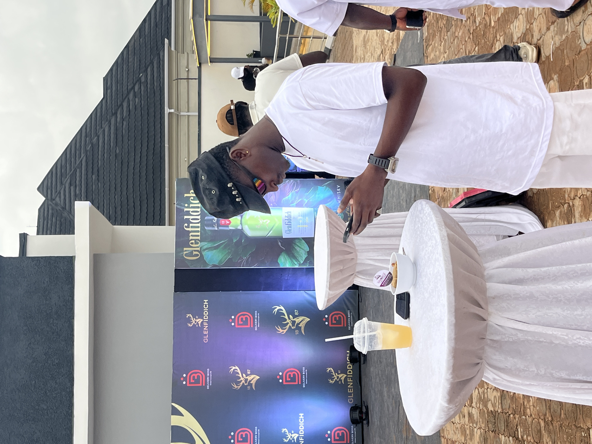 A young Black man in a white t-shirt and white pants stands at an outdoor event, looking down at his phone. He wears a black cap, sunglasses with rainbow-tinted lenses, and a watch. In front of him is a round white table covered with a patterned cloth, holding a plastic cup with a yellow beverage and straw, a small white bowl with snacks, and a black smartphone. Behind him, a backdrop features multiple logos, including "DELIVERY BOSS" and "GLENFIDDICH," along with images of a stag. Other people are visible in the background. The scene appears to be in an urban setting, possibly Bar Gada, Nigeria, with modern architecture and overcast skies.