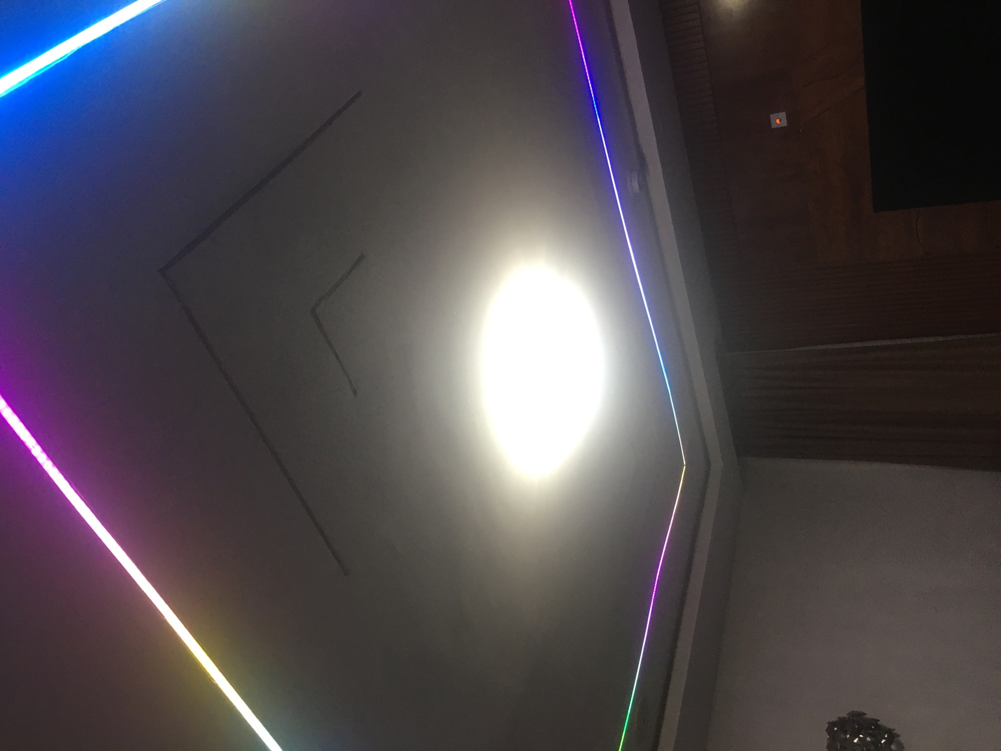 The image shows an interior view of a room with a light source dominating the center of the ceiling. RGB LED strips are positioned along the edges of the ceiling, creating a colorful border. The ceiling appears to have a geometric design. The upper right corner reveals a section of a wooden surface. The lower right corner shows a section of a wall. The photograph was presumably taken indoors. Location in Jos, Nigeria, is not discernible from the image.