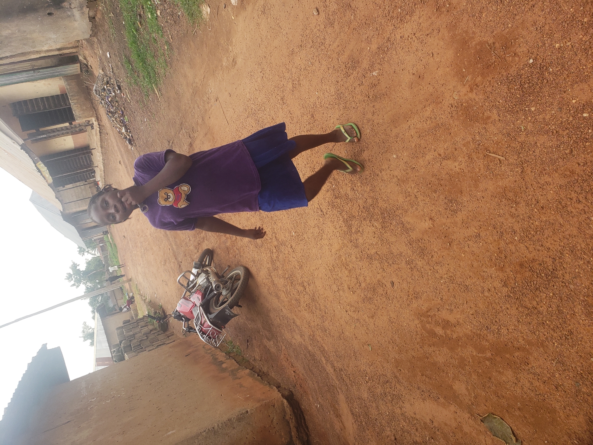In the city of Kachia, Nigeria, a young girl stands in the foreground on a dirt road. She is wearing a purple t-shirt with a teddy bear graphic and a blue skirt, along with green flip-flops. Her right hand is raised to her chin as she looks towards the camera. Behind her, a red motorcycle is parked on its side on the dusty road. In the background, there are buildings with corrugated metal roofs and a few other individuals are visible further down the road. The environment appears to be a rural or semi-urban area with unpaved roads and simple structures.