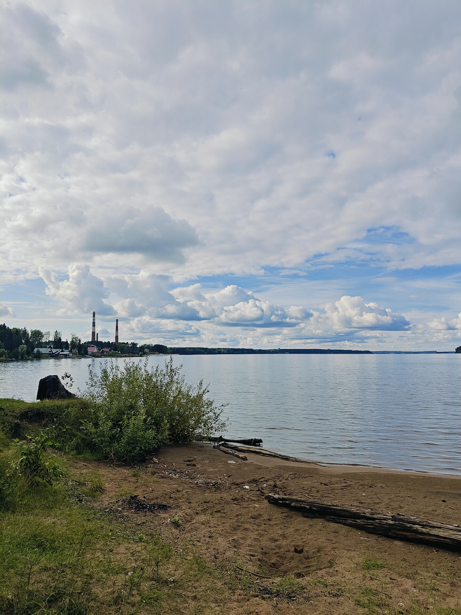 The image depicts a serene natural landscape on a cloudy day, likely near Chaikovsky, Russia, given the presence of industrial chimneys that are characteristic of the city's power plants. In the foreground, a sandy beach slopes down to a calm body of water, possibly the Kama River or a reservoir. Scattered on the beach are pieces of driftwood, and a dense green bush grows near the water's edge. Beyond the beach, on the left side of the image, is a grassy embankment with a large, dark, rocky object. In the distance, across the water, a line of trees marks the horizon, and two tall industrial chimneys, one red and white striped and the other solid red, stand prominently against the sky. The sky is filled with large, white cumulus clouds interspersed with patches of blue. The overall atmosphere is tranquil and reflects a natural setting with a touch of industrial presence.
