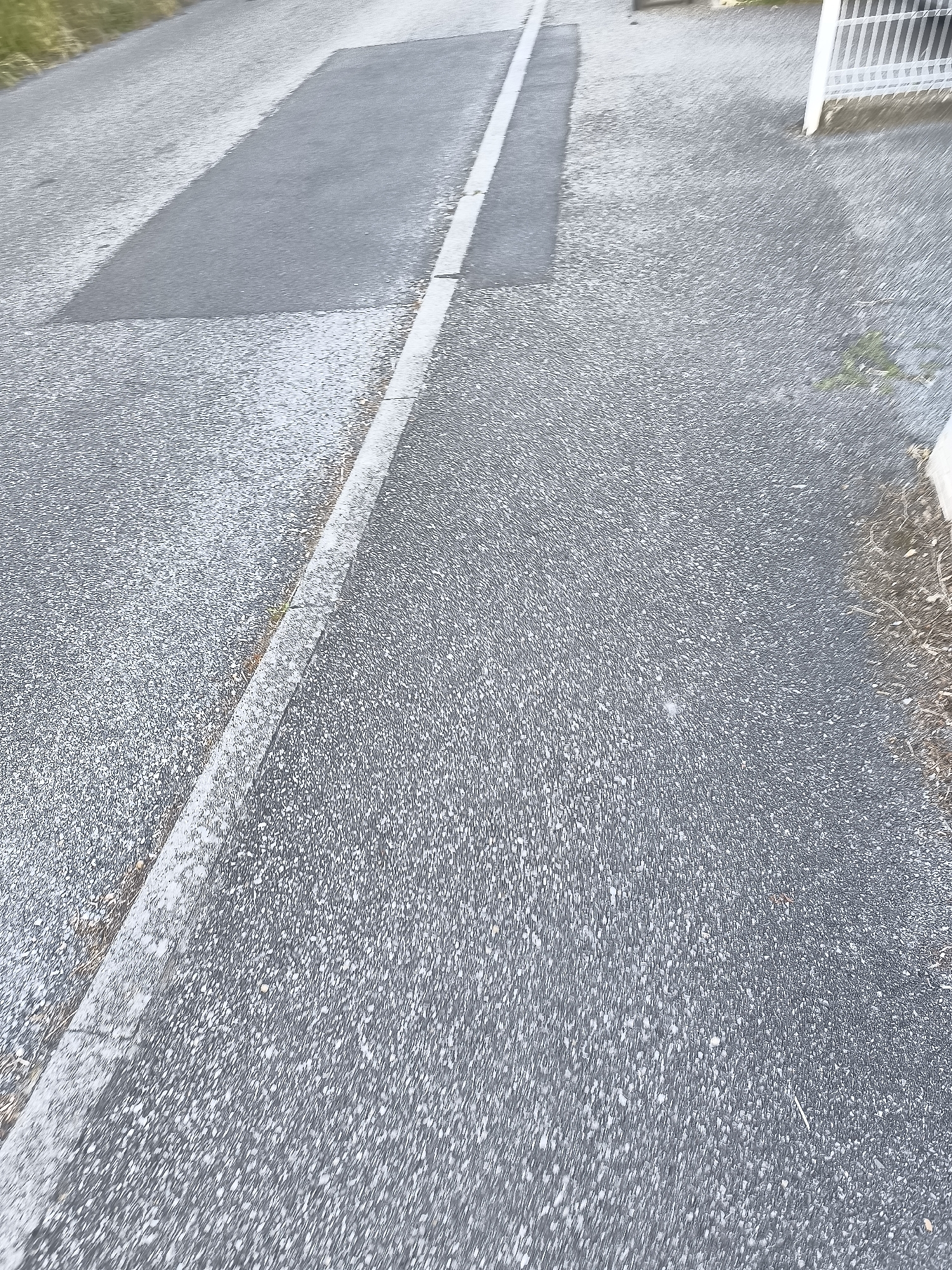 The image shows a paved surface in Pélussin, France. The pavement is gray and has a rough texture. A rectangular patch of darker gray pavement is visible on the left side of the image. A white line runs along the edge of the darker pavement, dividing it from the lighter gray pavement. To the right, there's a section where some debris and possibly vegetation are present, along with a glimpse of a white fence. No people are visible in the frame.