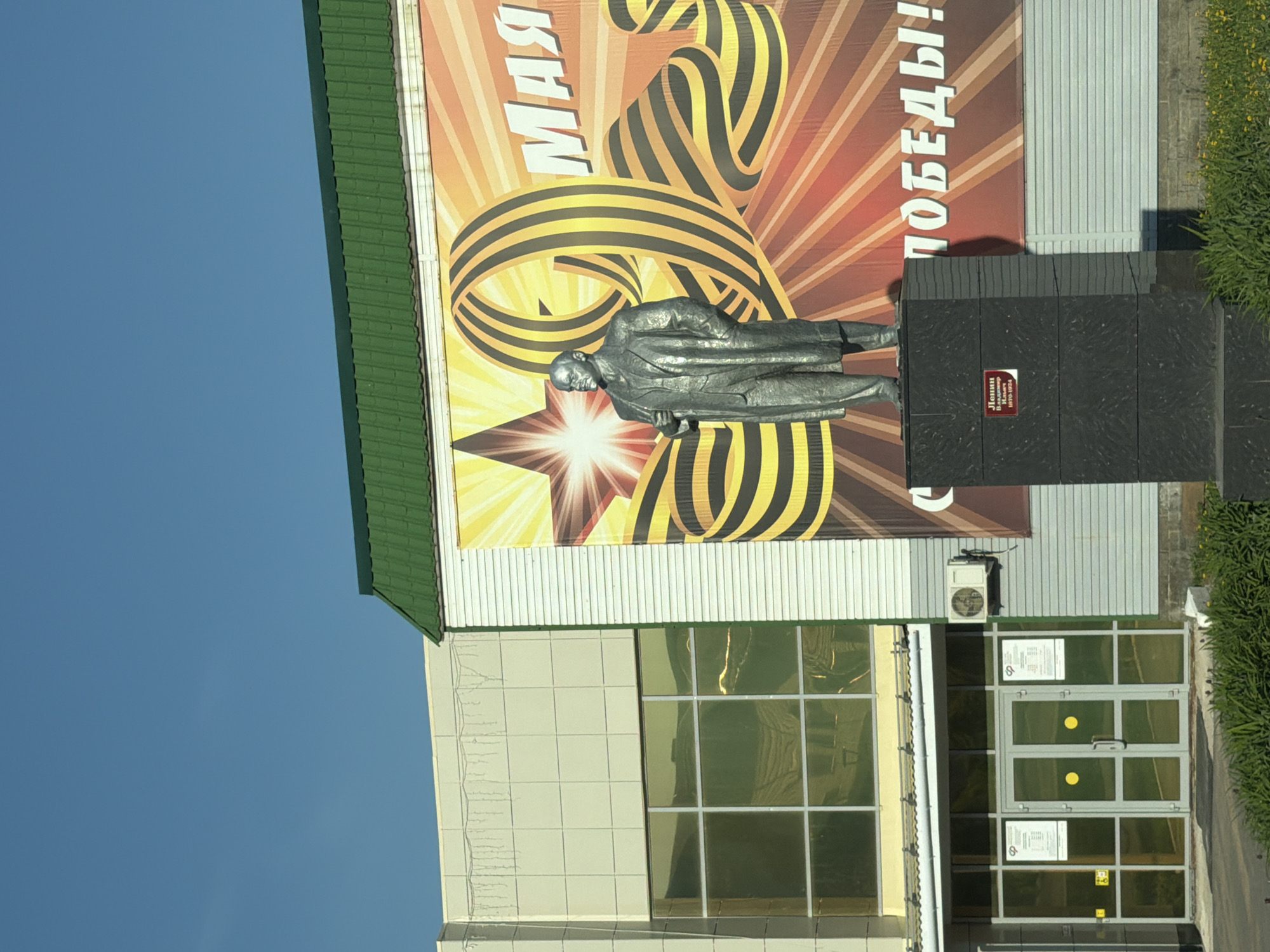 The image shows a statue of Vladimir Lenin in Agryz, Russia. The statue stands in front of a building adorned with a banner celebrating Victory Day on May 9th. The banner features the Saint George ribbon. The building has a green roof, tiled facade, and glass doors. An air conditioning unit is visible near the entrance. Vegetation is growing on the side of the building. The sky is clear and blue.
