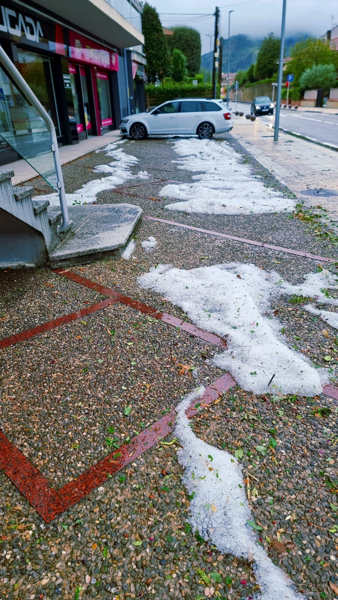 The image depicts a street in Sant Joan Les Fonts, Spain, after a hailstorm. Piles of hailstones, mixed with leaves, are scattered across the pavement and sidewalk in front of a building with glass storefronts. A white station wagon is parked on the side of the road, partially covered by hailstones. Further down the street, another car is visible. The sky is overcast, suggesting continued inclement weather. The ground is a mixture of gravel and red brick lines. A set of concrete stairs with a metal railing leads up to the building's entrance.
