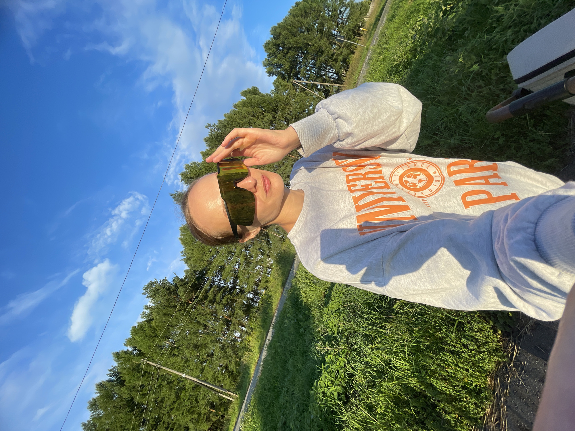 A young woman with her hand near her large, reflective sunglasses is standing outdoors in a grassy area next to a road. She is wearing a light gray sweatshirt with orange lettering that reads "UNIVERSITY OF SCIENCE" and "PARIS" with a globe emblem. The background consists of green trees and a blue sky with scattered clouds. The scene appears to be in the village of Podshivalovo, Russia.