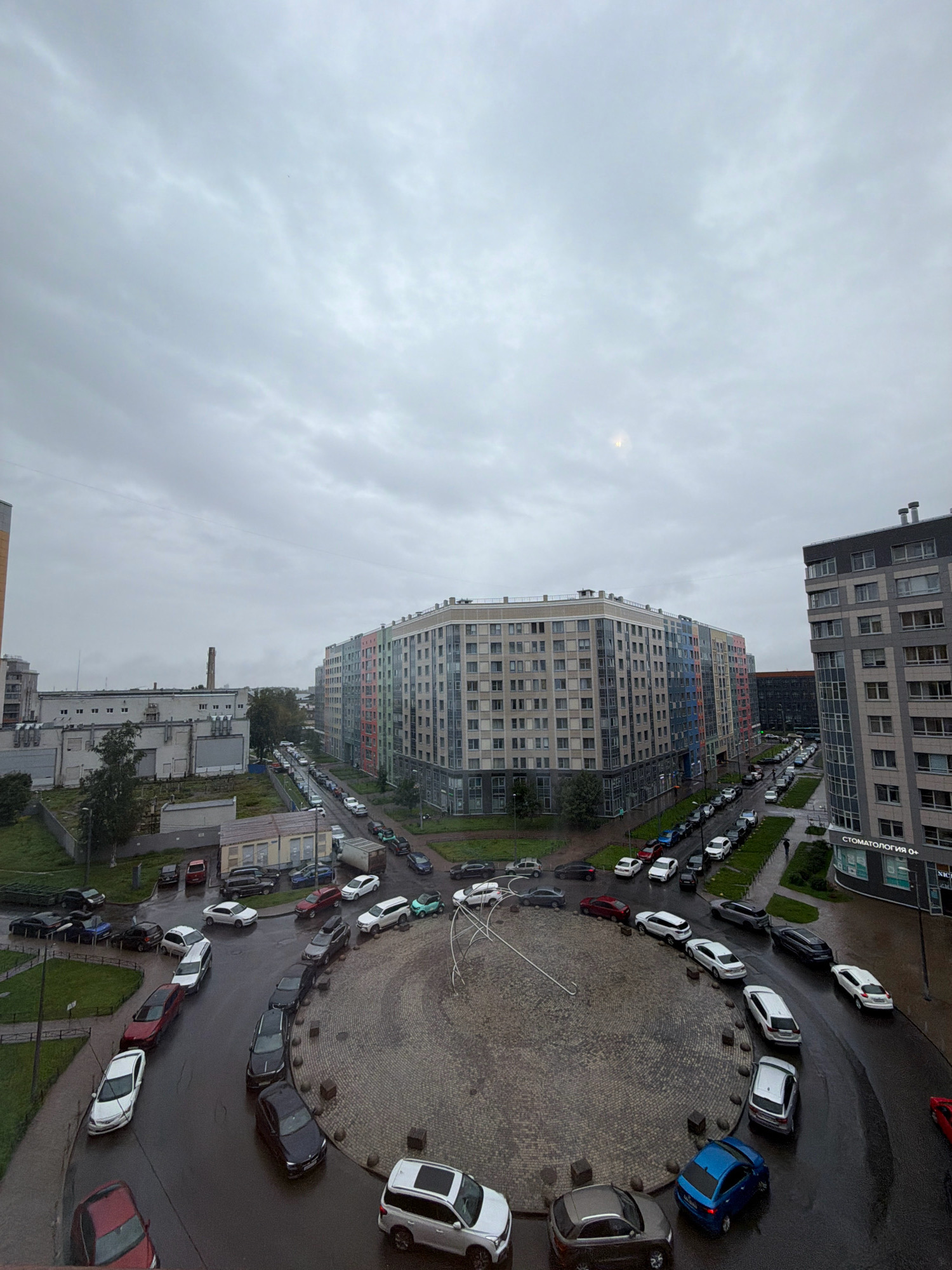 The image depicts a cloudy day in a residential area of Saint Petersburg, Russia, specifically the Novoorlovsky residential complex. The scene is dominated by a large, multi-colored apartment building that curves around a circular paved area. Numerous cars are parked and driving in and around this circular space, as well as along the adjacent roads. Several other buildings, including a taller, grey apartment building with a "Стоматология" (Dentistry) sign, are visible. The sky is overcast with a dull grey hue. The ground appears wet, suggesting recent rain. There are no visible people clearly engaged in any specific actions. The overall atmosphere is that of a typical urban environment under cloudy skies.
