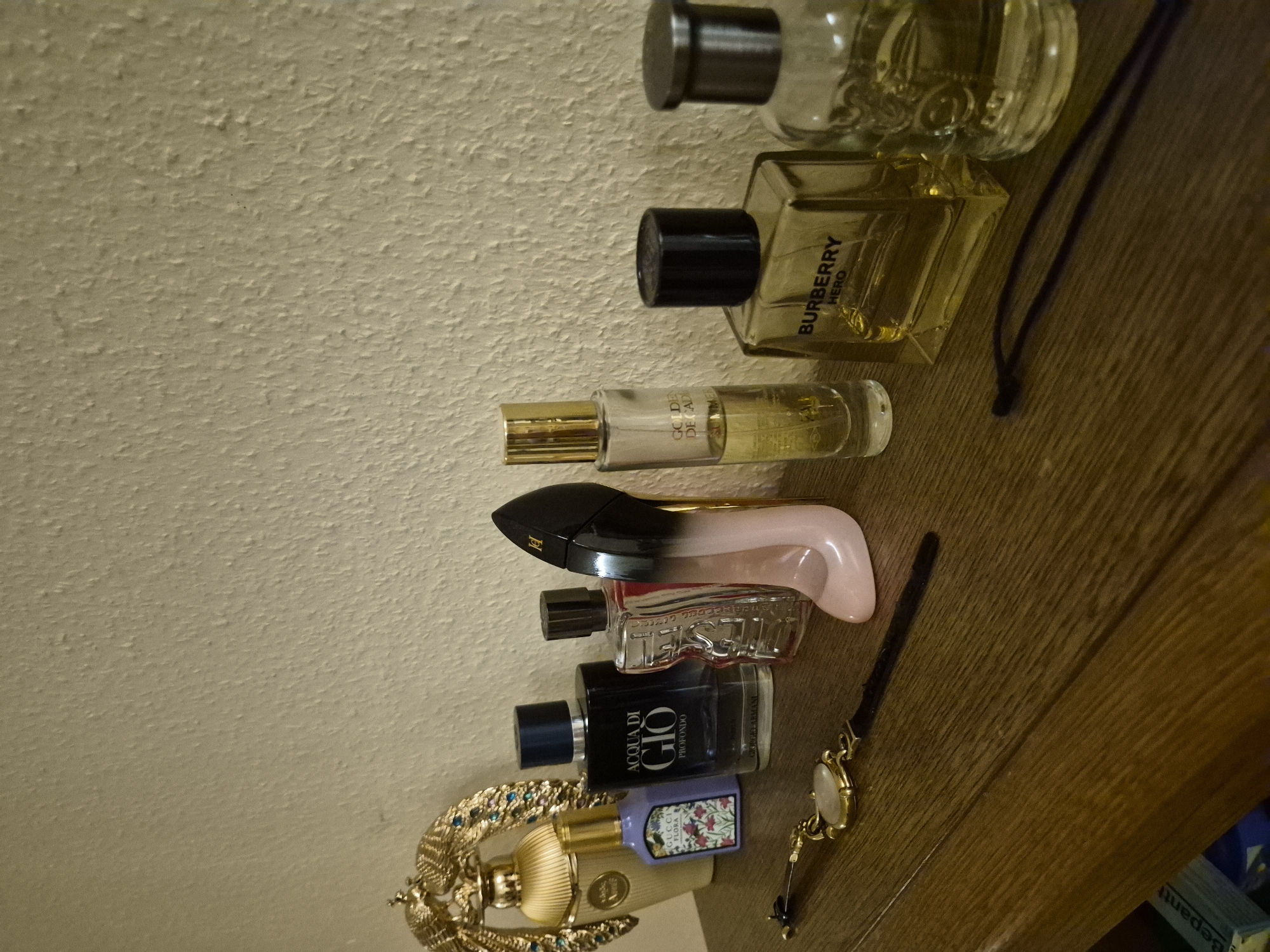 A high-angle shot shows a collection of perfume bottles arranged vertically on a textured, beige surface next to a wooden surface. From top to bottom, the bottles are: a clear glass bottle with a dark cap, a square-shaped gold-toned glass bottle with a black cap and the text "BURBERRY HERO" printed on it, a slender clear glass bottle with a gold cap and text that appears to say "GOLDEN DECAF," a black and pink bottle with an unusual shape, a clear glass bottle with red liquid and a black cap, a black bottle with a dark blue cap and text that reads "ACQUA DI GIO PROFONDO GIORGIO ARMANI," and a small purple bottle with a floral label. Below these are a gold-toned decorative object, possibly a trophy or a clasp, and a gold-toned wristwatch with a light-colored face and a black band. The objects are placed on a light-colored, textured surface that transitions to a darker, wood-grained surface. The lighting appears to be artificial, casting shadows. No people are visible, and the scene seems to be a still-life arrangement of personal items.