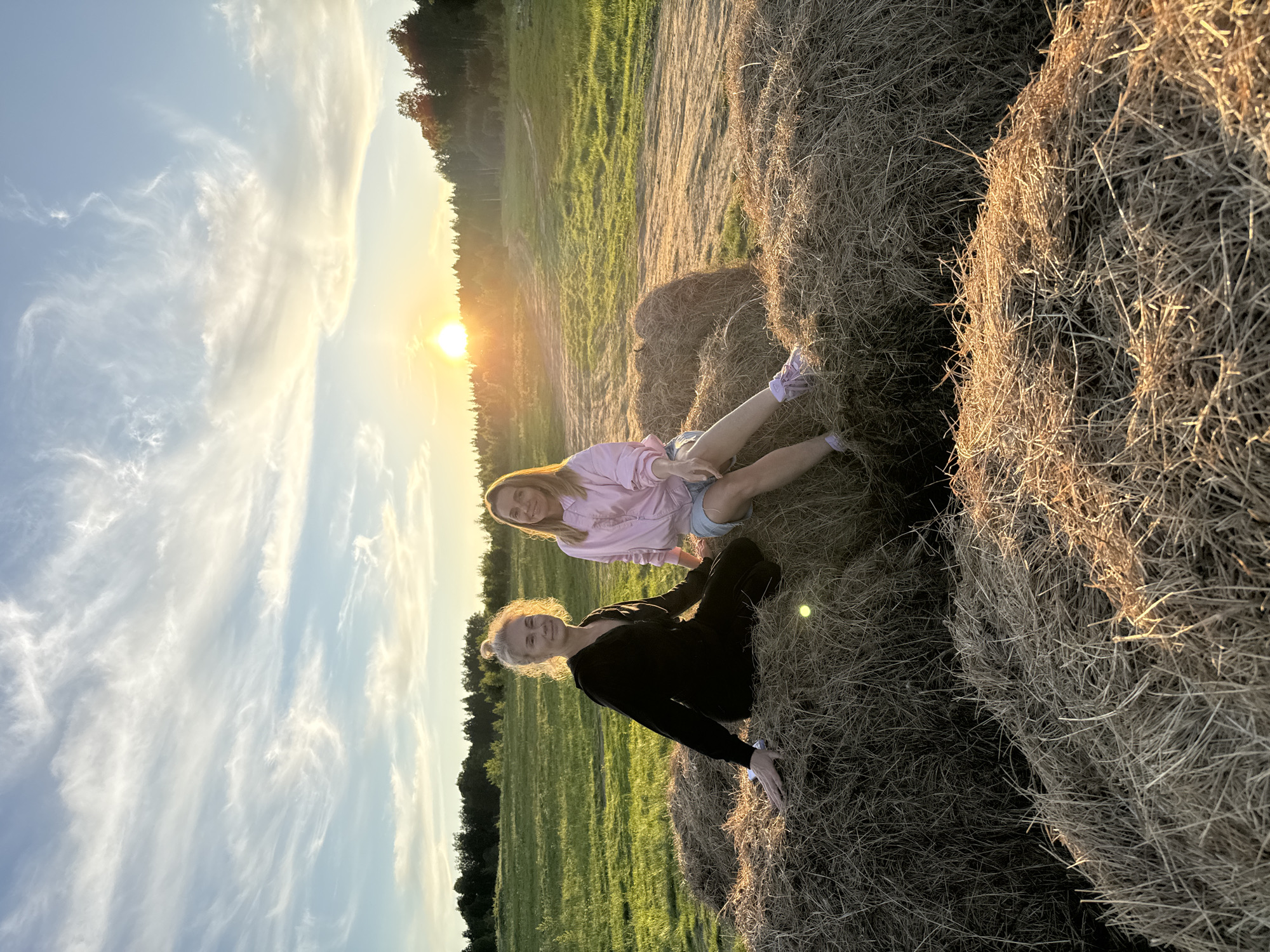 Two women are seated on large hay bales in a golden-hour field, bathed in the warm light of a setting sun. The image captures a serene outdoor scene, likely in a rural or agricultural area near Стеклянный, Russia.
One woman, positioned in the foreground to the left, has blonde hair tied back and is wearing a black long-sleeved top and dark pants. She is smiling gently at the camera. Behind and to her right, a second woman with long blonde hair is dressed in a pink long-sleeved top and denim shorts, also smiling towards the viewer. Both appear relaxed and joyful.
The hay bales they sit upon are textured and appear to be made of dried grass or straw, strongly illuminated by the low sun. Beyond the hay bales, a vast green field stretches into the distance, bordered by a line of trees on the horizon. The sky is a prominent feature, displaying a bright, setting sun on the right, which casts a golden glow and produces lens flare. The sky transitions from a soft blue at the top to vibrant oranges and yellows near the sun, with scattered wispy clouds. The overall ambiance is peaceful and pleasant. No text is visible in the image.
