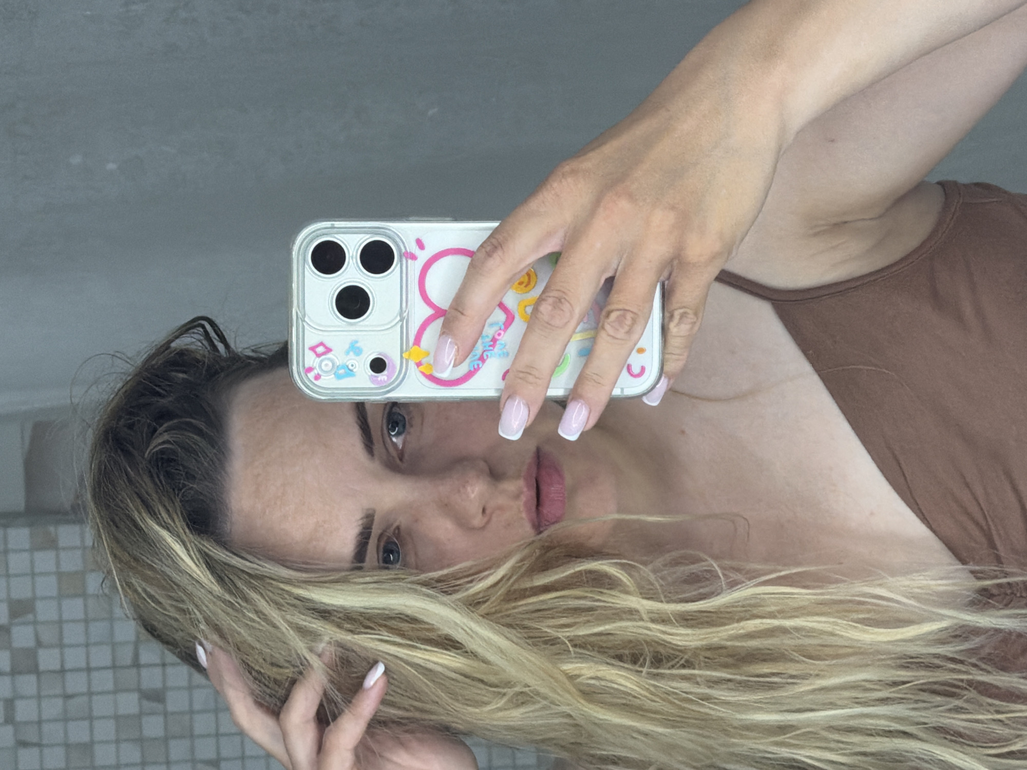 A woman with blonde hair is taking a selfie in front of a mirror. She is holding a phone with a white case decorated with colorful cartoon figures. The background appears to be a bathroom or a room with tiled walls. The woman is wearing a brown tank top. There is no indication of the location being Votkinsk, Russia.