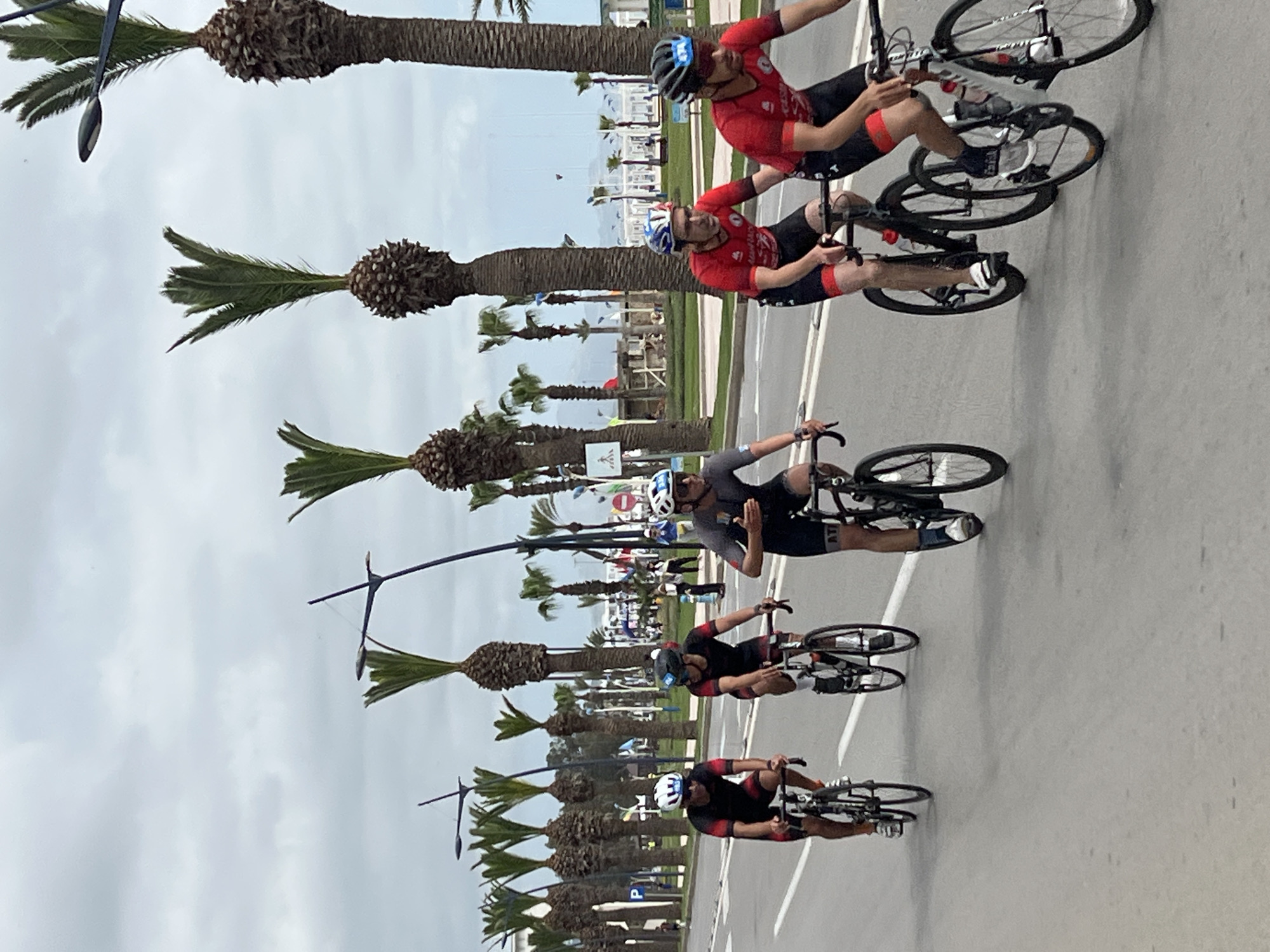 The image captures a group of cyclists in M'Diq, Morocco, riding along a road lined with palm trees. The sky is overcast. Five cyclists are visible, each wearing helmets and athletic gear. Some cyclists wear red jerseys with black shorts, while one wears a grey top with black shorts. The cyclists are spread out, suggesting a race or training session. The road is grey and marked with white lines. In the background, buildings and structures are visible, indicating an urban setting near a coastline. The palm trees are evenly spaced and lined up along the roadside, suggesting a deliberate landscaping design. Streetlights with decorative arms are interspersed among the palm trees.