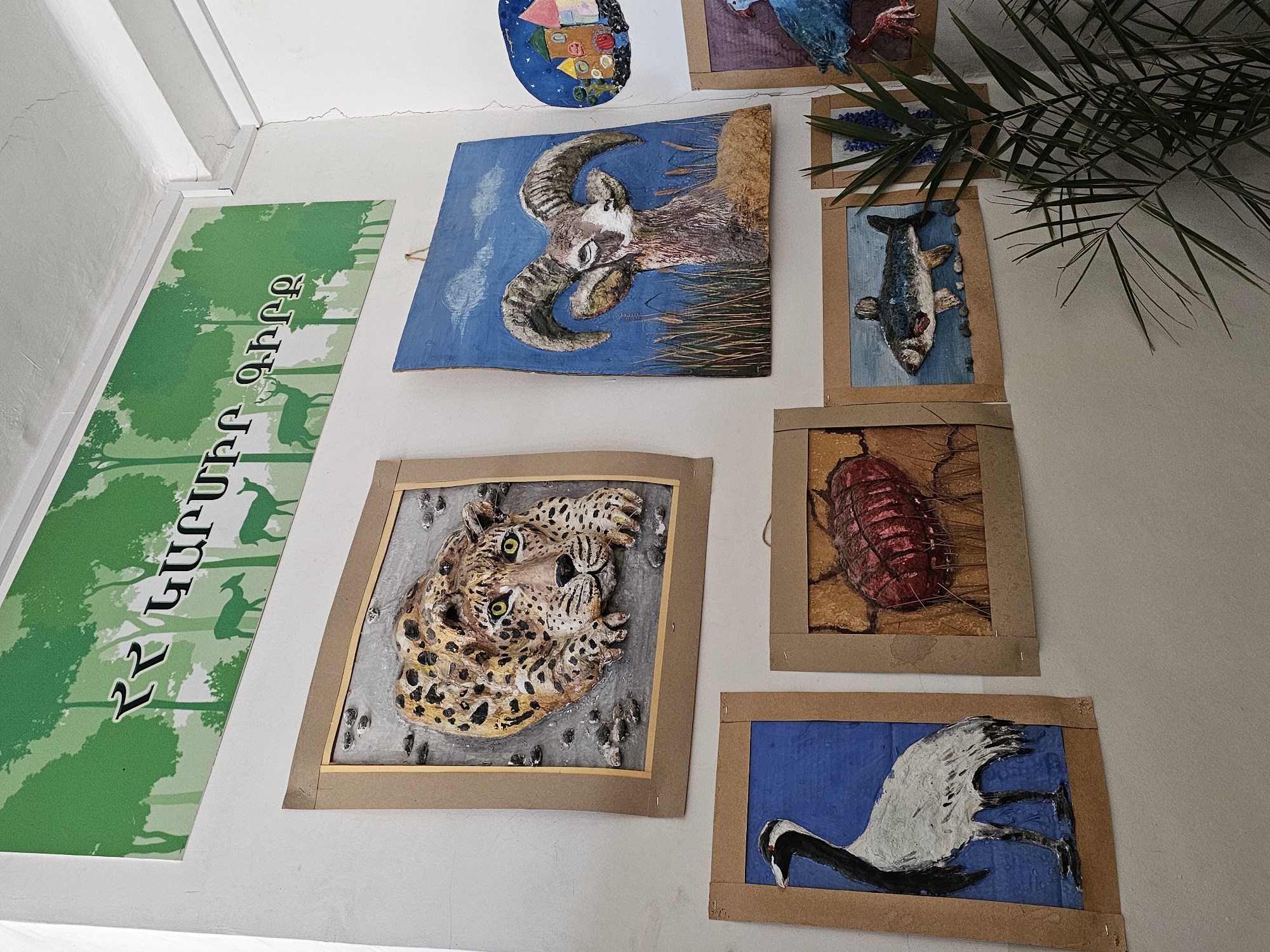 The image shows a wall decorated with several framed artworks. The artworks depict various animals including a ram, a leopard, a fish, an insect, and a bird. There is also a green banner with text in Armenian, and a depiction of trees and deer. A leafy plant is situated to the right of the framed artworks. Based on the subject matter of the artwork and the presence of Armenian text, it is likely that the location is within the city of Yerevan, Armenia.