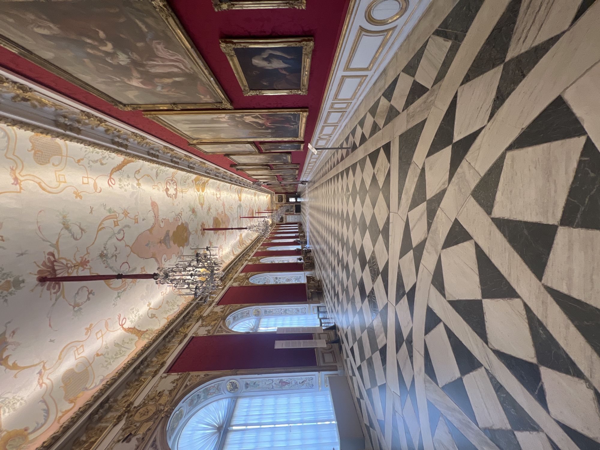The image depicts the Great Hall of the New Schleißheim Palace in Oberschleißheim, Germany. The hall is a long, ornate corridor with a black and white checkered marble floor. The walls are a deep red and adorned with numerous framed paintings. The ceiling is intricately decorated with frescoes and gold trim, and several crystal chandeliers hang from it. Large arched windows with white translucent curtains line the left side of the hall, letting in natural light. The hall appears to be empty of people, with the exception of a few figures visible in the distance at the far end of the corridor. The overall impression is one of grandeur and historical significance.