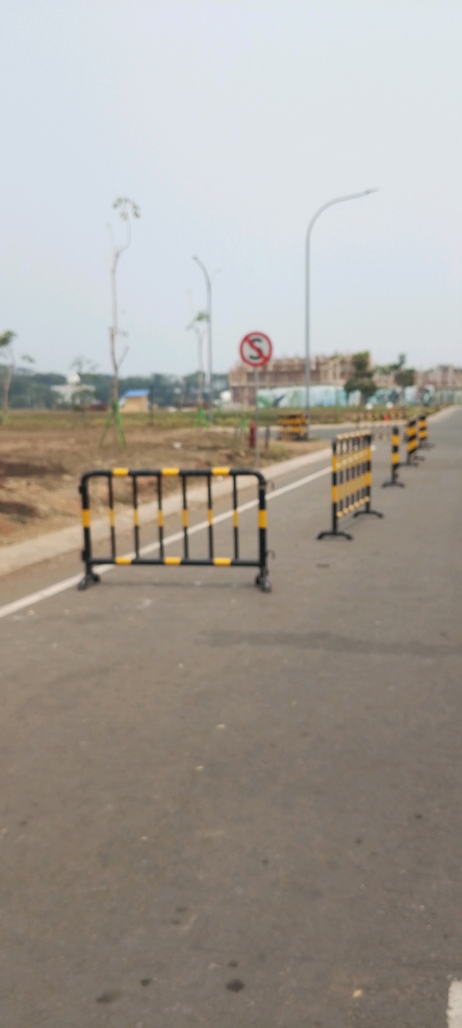 A black and yellow striped metal barricade is positioned on an asphalt road. The barricade is oriented horizontally and is the closest object to the viewer. In the background, a white line marks the edge of the road. Further down the road, more barricades of the same design are visible, receding into the distance. A red and white circular sign with a curved arrow, indicating a prohibition, is also visible in the background. The landscape beyond the road appears to be under construction, with buildings in various stages of completion. The sky is overcast. The location is Gunung Putri, Indonesia.
