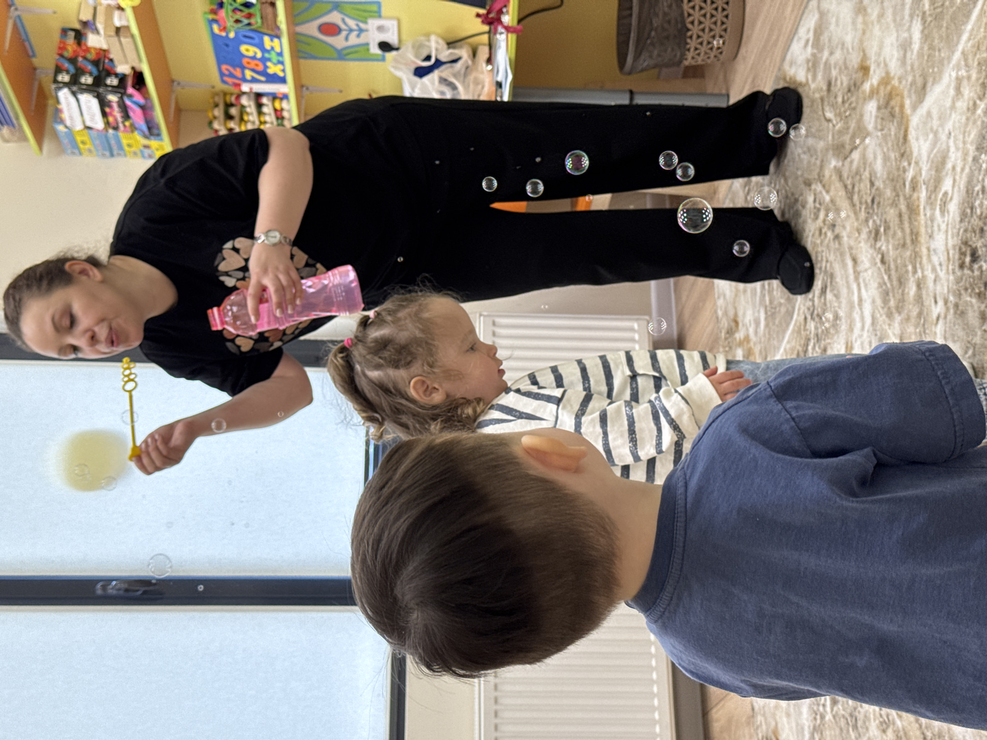 The image depicts an engaging indoor scene in what appears to be a daycare, playroom, or classroom, located in Izhevsk, Russia. The setting is bright with natural light, suggesting it is daytime.
An adult female, positioned on the left, is actively blowing bubbles for two young children. She has brown hair, wears a black t-shirt adorned with a subtle heart pattern, black pants, and a wristwatch on her left wrist. She holds a yellow bubble wand in her left hand, from which bubbles are emerging, and a pink bottle, presumably filled with bubble solution, in her right hand. Her expression appears gentle and focused on the activity.
In the middle-right, a toddler girl with light brown curly hair tied in pigtails is seen in profile, attentively watching the bubbles. She is wearing a white and dark striped long-sleeve shirt. In the foreground, on the right, a young boy with short brown hair is seen from behind, wearing a blue t-shirt, also looking towards the bubble activity. Numerous iridescent bubbles are suspended in the air around the subjects, adding a playful element to the scene.
The background on the left features shelves stocked with colorful educational or play items, including blocks with visible numbers ("12", "7890") and letters ("X+H"). A large window in the mid-left lets in natural light, with a white radiator visible beneath it. The floor is a light-colored surface with a marble-like pattern. The scene conveys a sense of joy, engagement, and focused attention from the children during a playful activity.