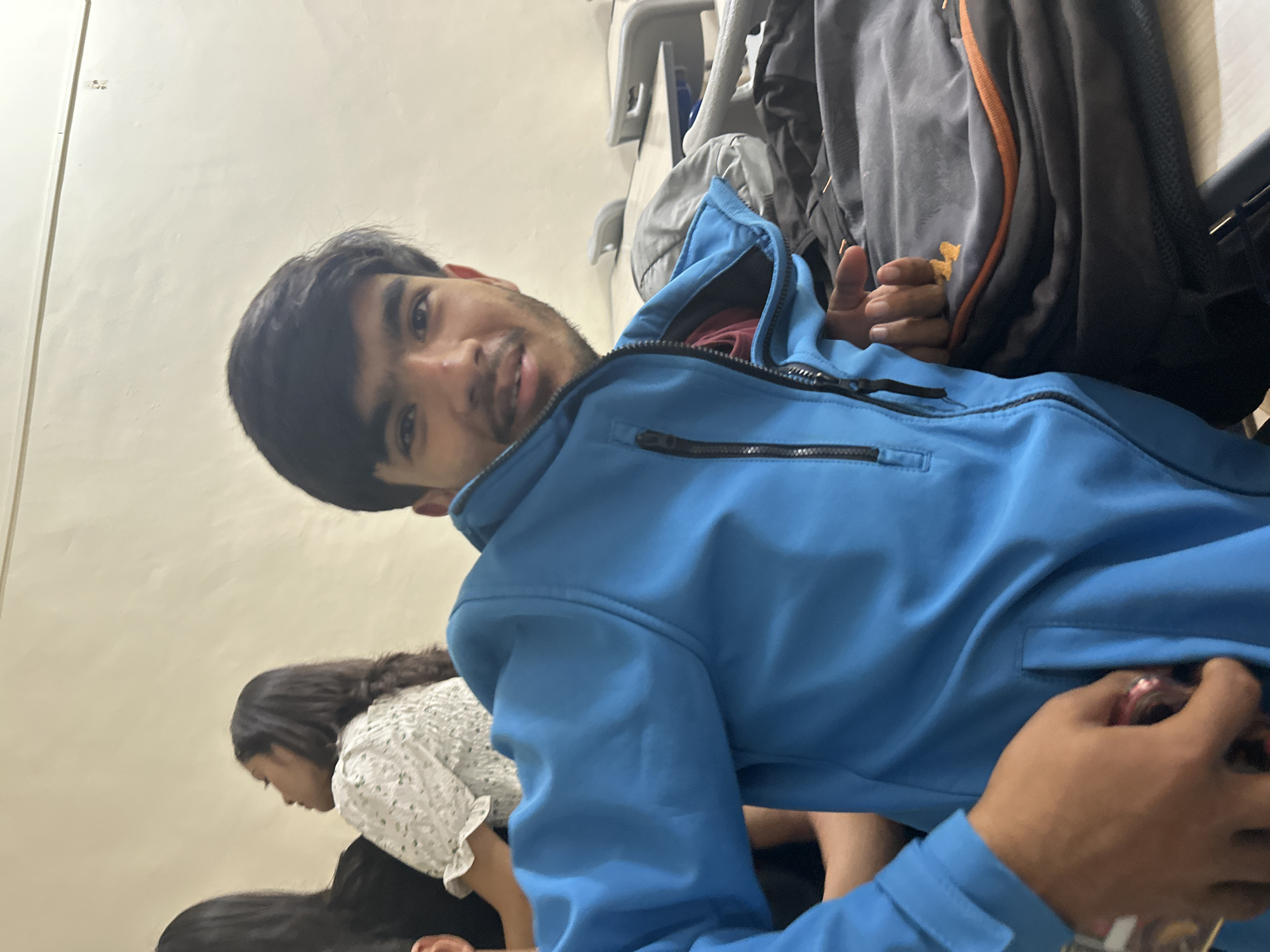 In an indoor setting, likely a classroom due to the presence of desks and chairs, a young man is the primary subject. He is wearing a bright blue zippered jacket over a burgundy garment, and his left hand is partially visible, holding a small, shiny object. Behind him, two young women are visible, one in a white patterned top and the other in a black top. The background consists of beige walls and a white ceiling with some smudges. A dark backpack with an orange trim lies on a desk in the foreground. The lighting suggests an indoor, possibly artificial, light source.
