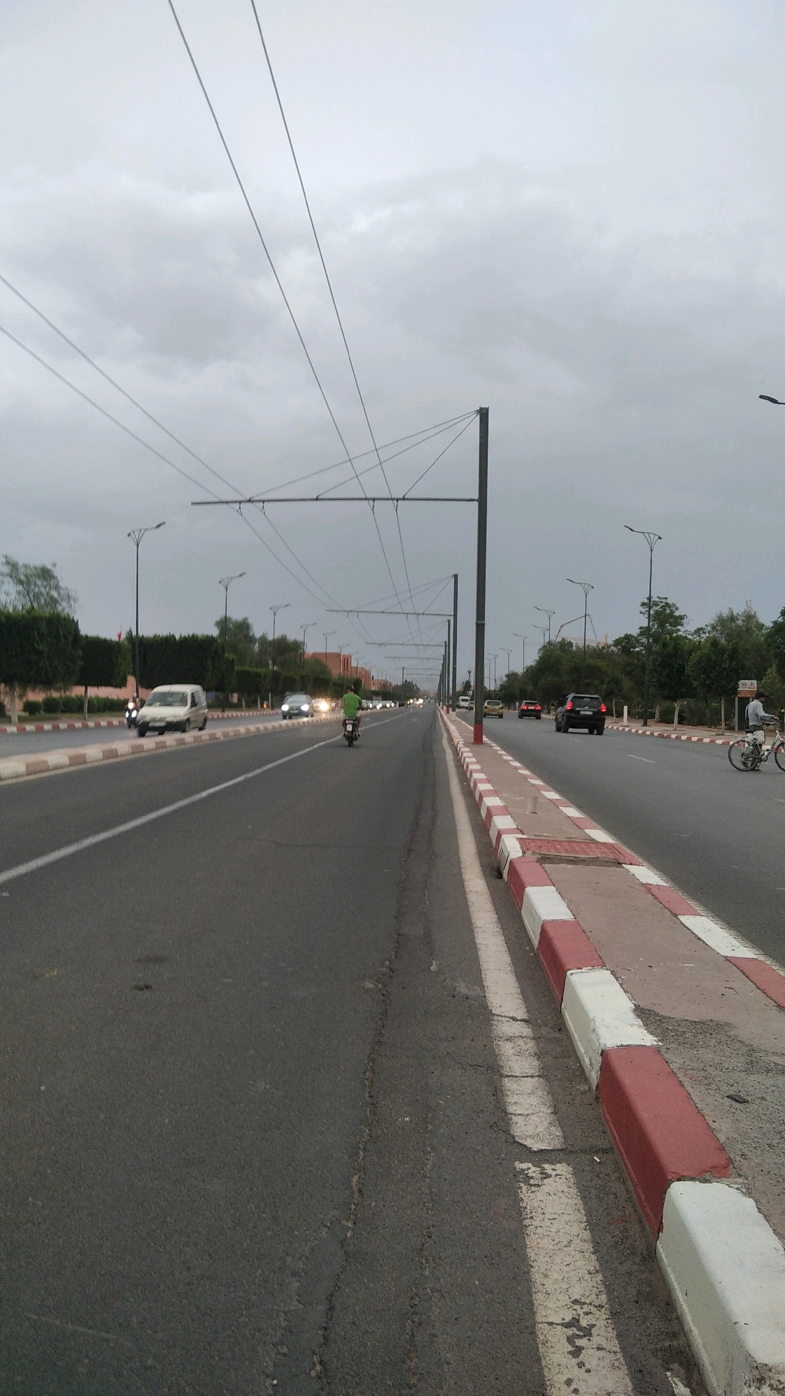 The image shows a wide road in an urban environment under an overcast sky. The road is asphalt with white lane markings. There is a concrete median strip with red and white colored borders. On the left side of the road, there is a sidewalk with a line of trees and some buildings visible in the distance. On the right side of the road, there is more vegetation and some signage. There are multiple overhead power lines supported by tall poles running along the road. Several vehicles are present, including cars, a white van, a motorcycle with a rider wearing a green shirt, and a bicycle with a rider. The scene appears to be a typical city street during daylight hours, with moderate traffic.