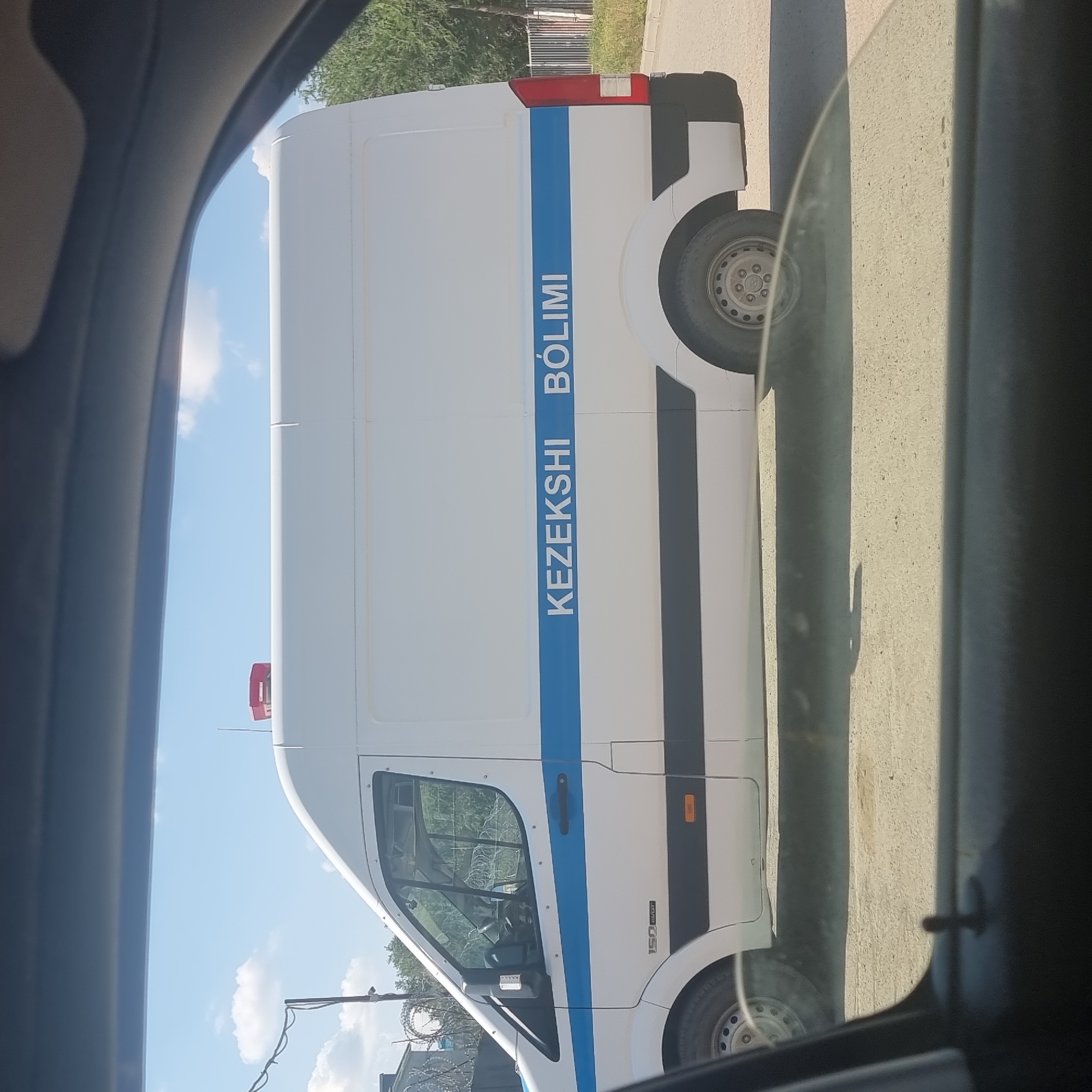 A white van with a blue stripe and the words "KEZEKSHI BÓLIMI" is parked outdoors. The van has a red light on its roof. The background includes trees and a fence. The image is taken from inside another vehicle, with a blurred foreground showing a car door handle. The van appears to be a police or official vehicle. The text "KEZEKSHI BÓLIMI" suggests it is from Aktobe, Kazakhstan, as this translates to "emergency department" or "ambulance service" in Kazakh, a language commonly spoken in the region.