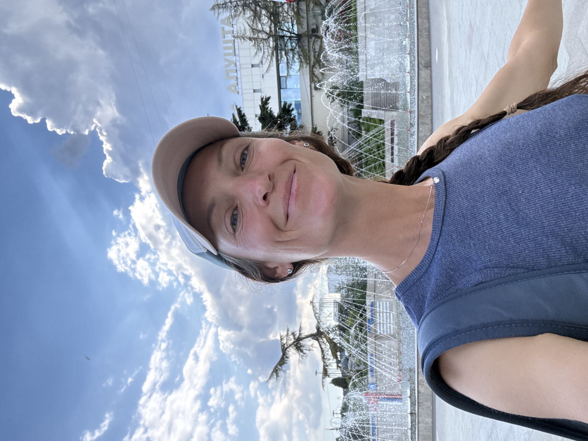 A woman is taking a selfie in front of a fountain and a building with the text "АЛУШТА" in Ukrainian. She is wearing a blue tank top and a baseball cap. The sky is blue with clouds.
