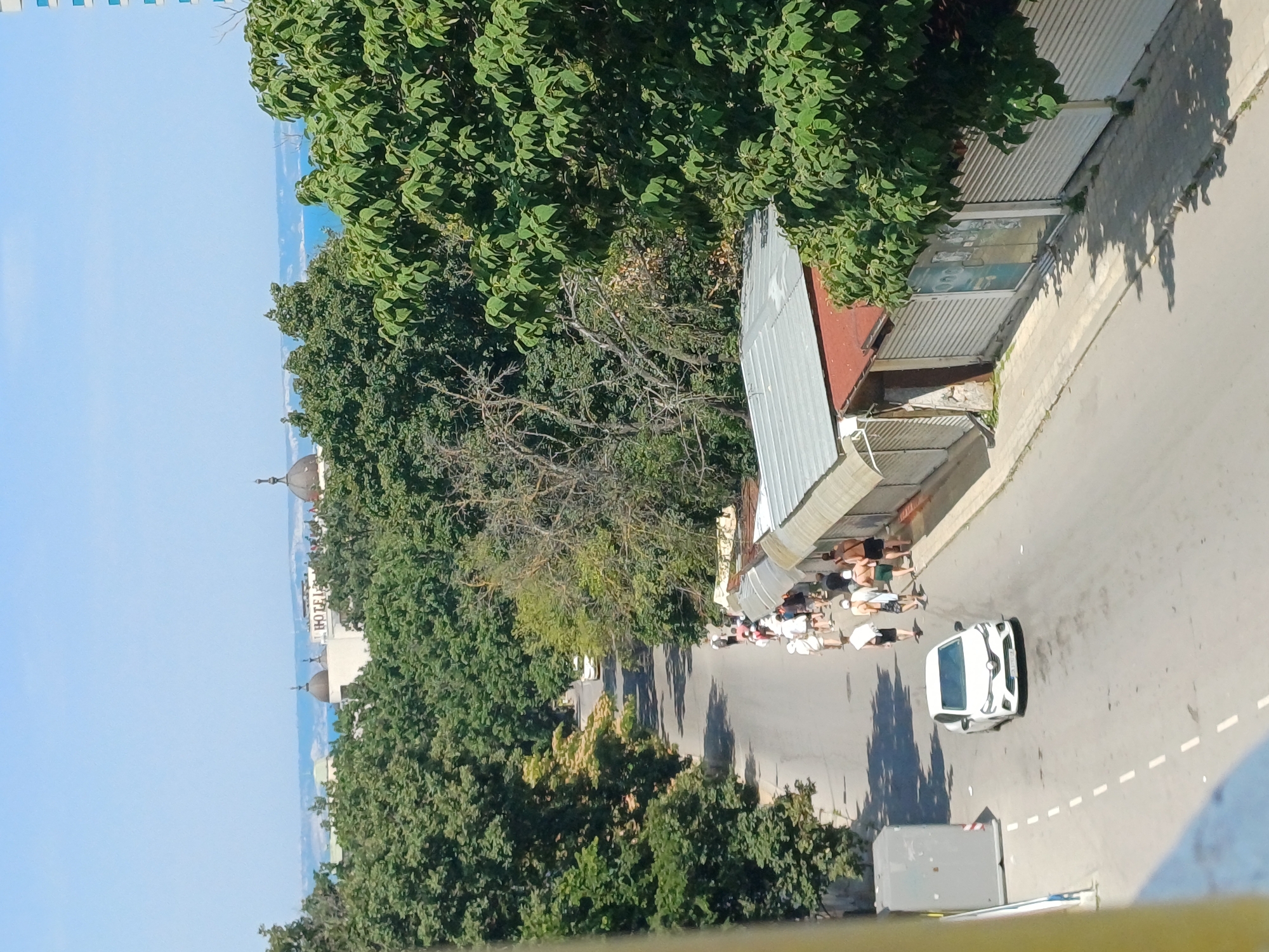In Varna, Bulgaria, on a sunny day, a white car drives down a paved road lined with shops and trees. A group of people walks along the sidewalk next to the shops, some wearing shorts and t-shirts, suggesting warm weather. In the background, beyond the trees, the blue sea is visible, and distant buildings with dome-like structures are seen, hinting at a coastal city setting. The shops appear to be closed or have their shutters down, with some displaying advertisements or signs. The overall scene captures a street with commercial activity and pedestrian traffic, set against a backdrop of lush greenery and the sea.